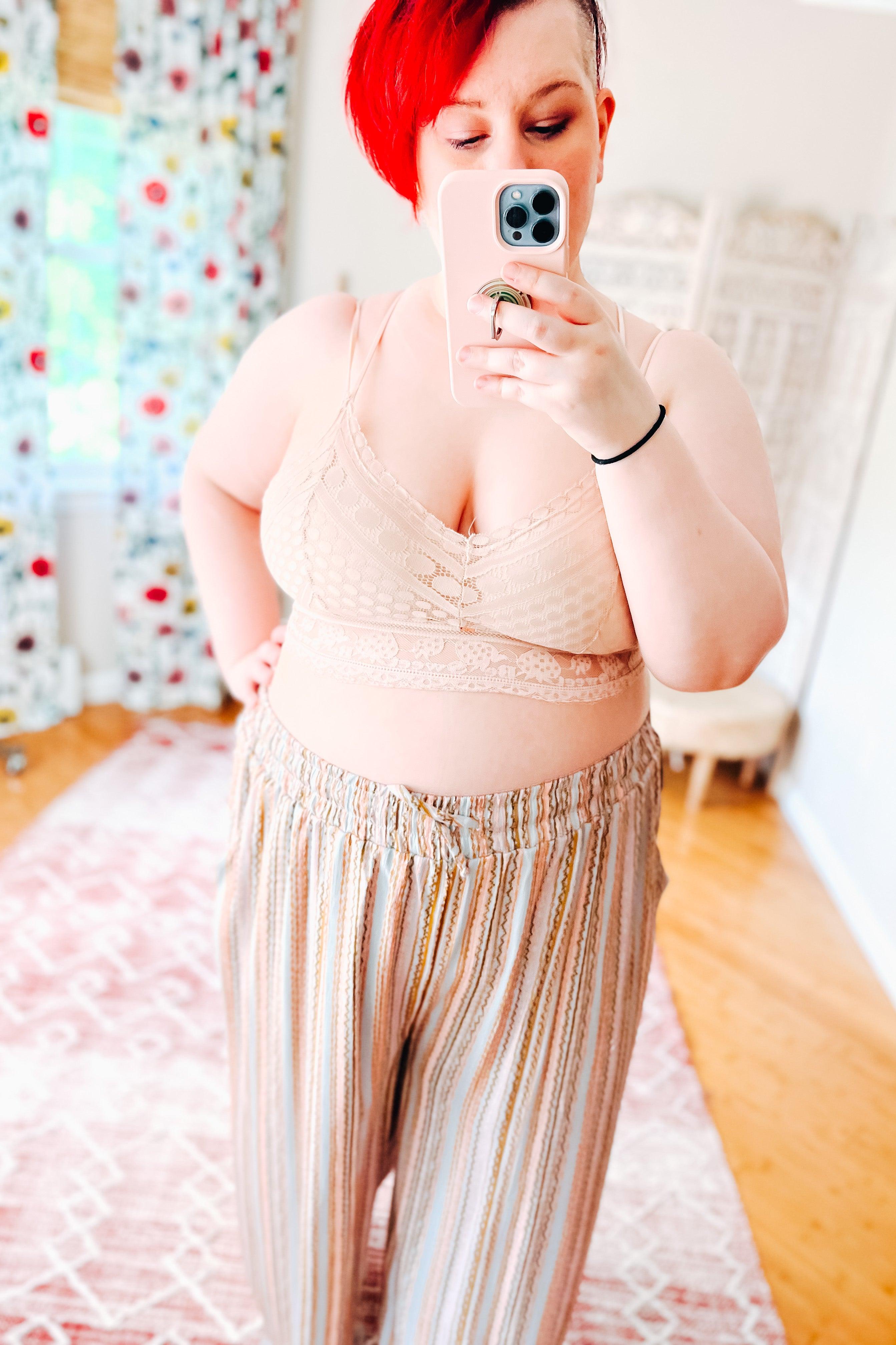 Woman with bright red hair wearing a blush lace bralette featuring detailed patterns paired with loose striped pants, taking a mirror selfie indoors.