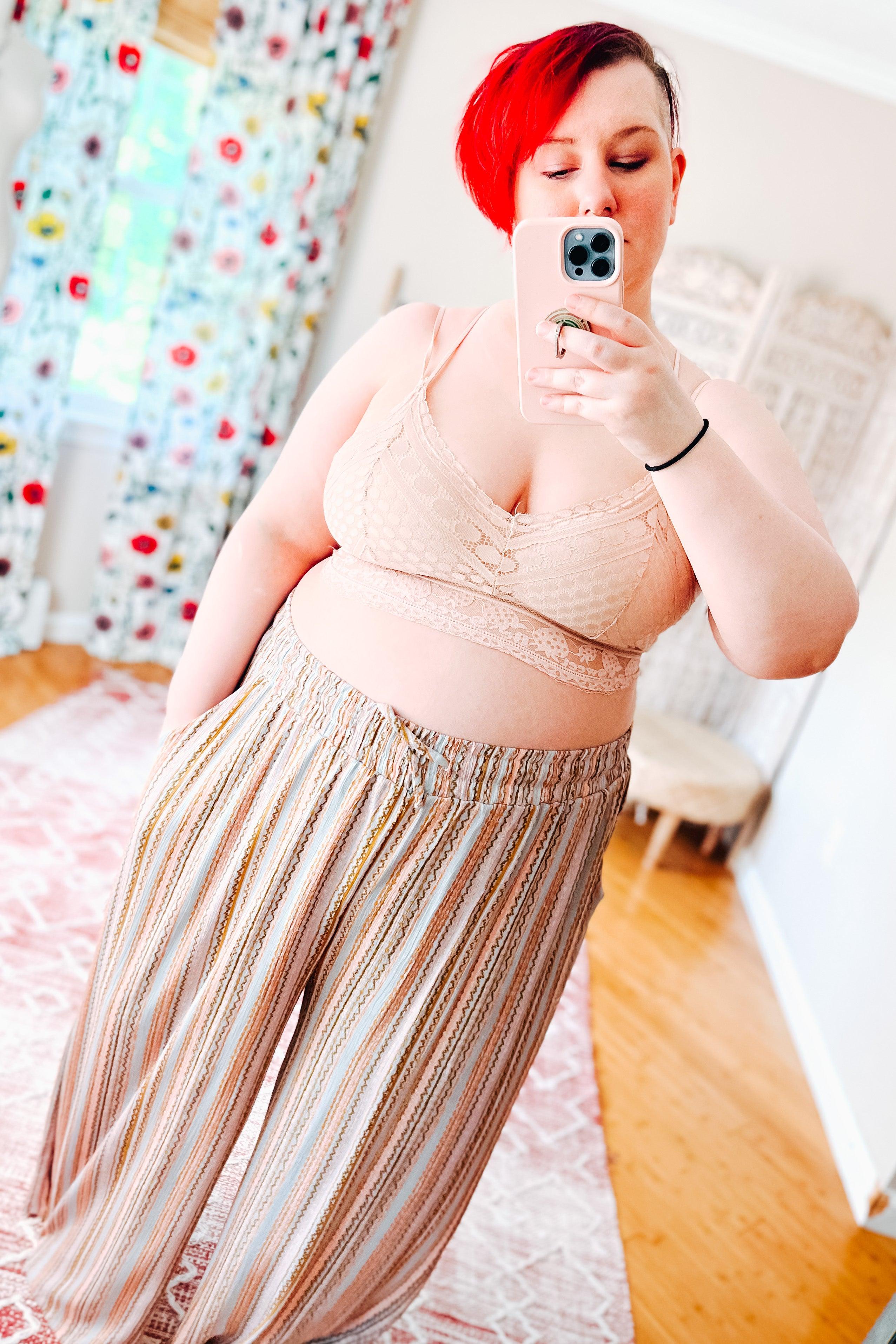 Woman with bright red hair wearing a blush lace bralette with delicate detailing and loose striped high-waisted pants, taking a mirror selfie indoors.