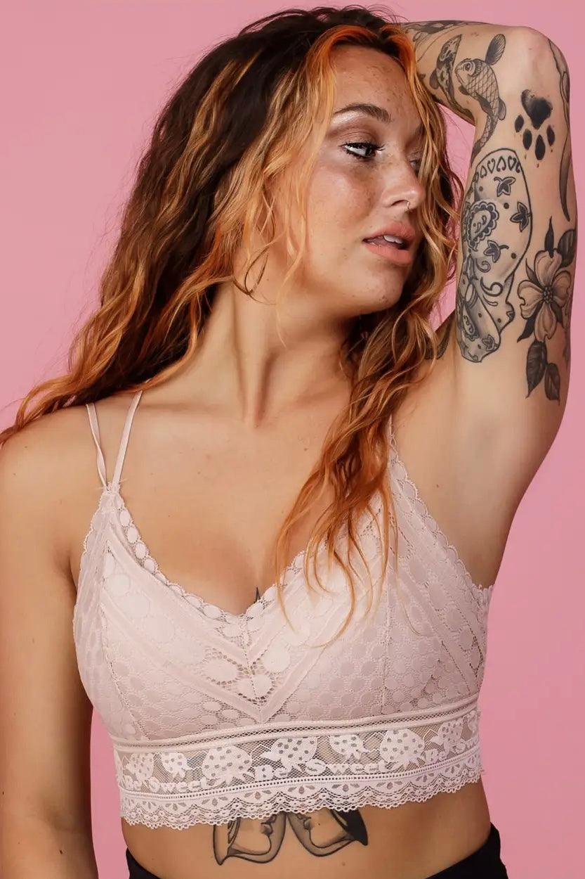 Close-up of a woman with tattoos wearing a blush lace bralette, posing against a pink background with relaxed expression and loose wavy hair.