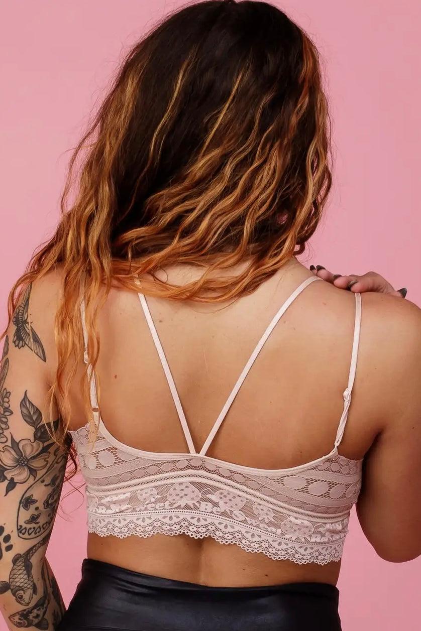 Rear view of a woman wearing a delicate blush lace bralette with thin straps and floral patterns against a soft pink background.