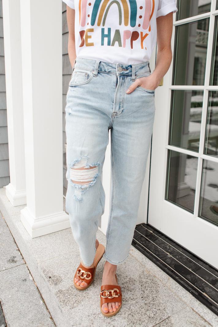 Light blue distressed jeans with ripped detailing on one knee, styled with casual sandals and a colorful graphic tee.