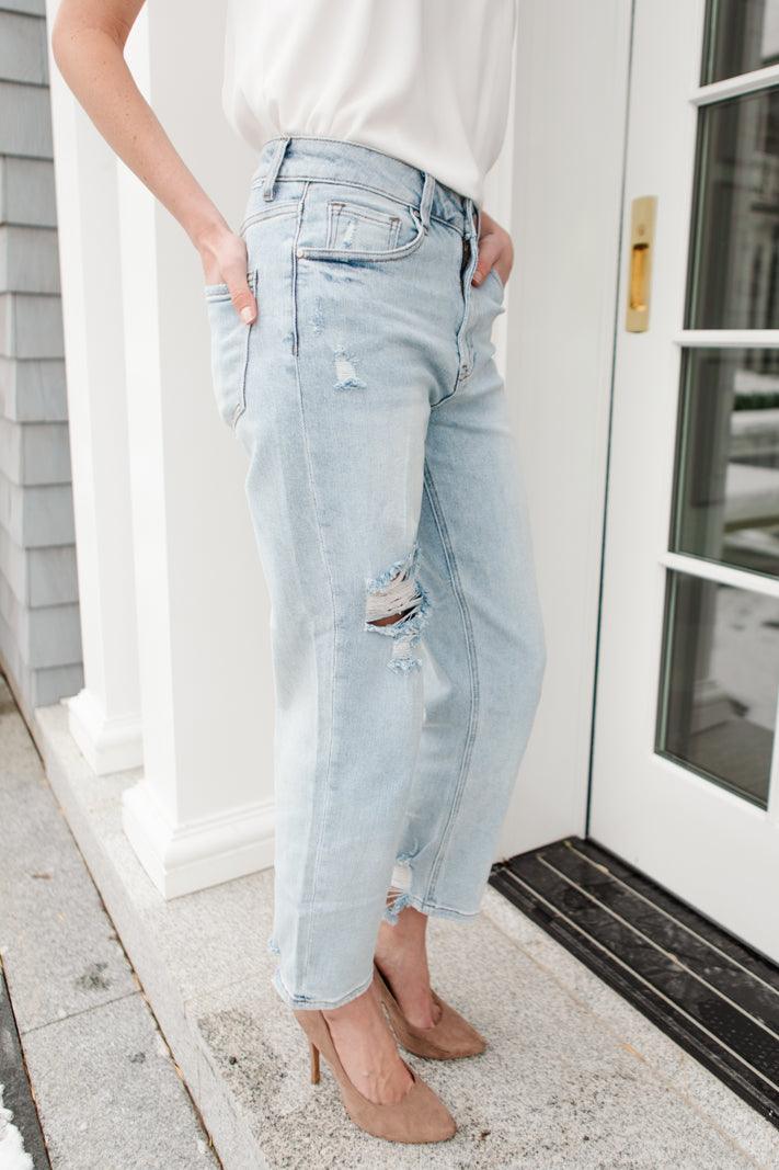 Light blue distressed jeans with ripped details on one thigh, styled with a white sleeveless top and beige pointed-toe heels outdoors by a door.