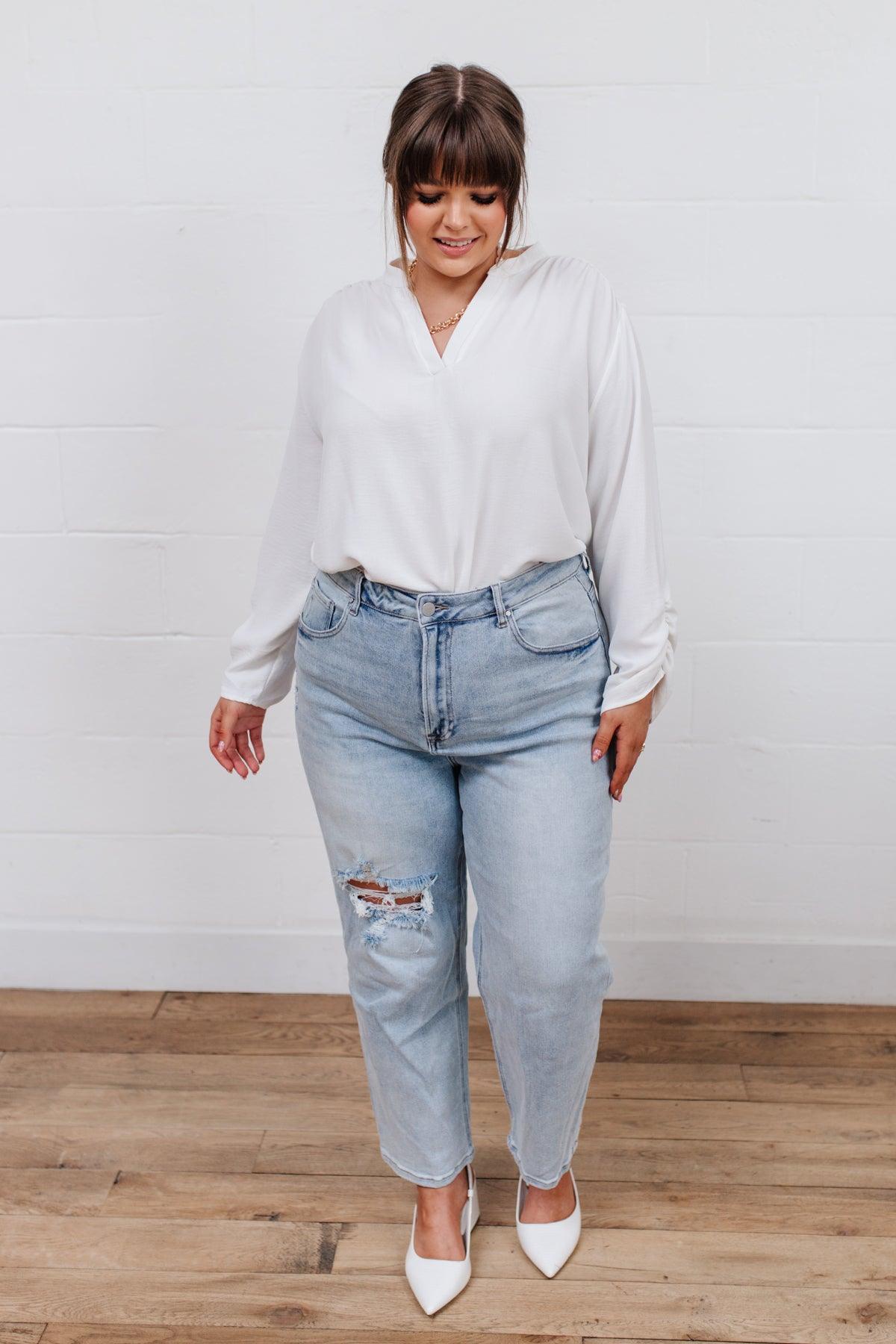 Woman wearing light blue jeans with a ripped knee detail styled with a loose white blouse and white pointed-toe heels standing indoors on wooden flooring