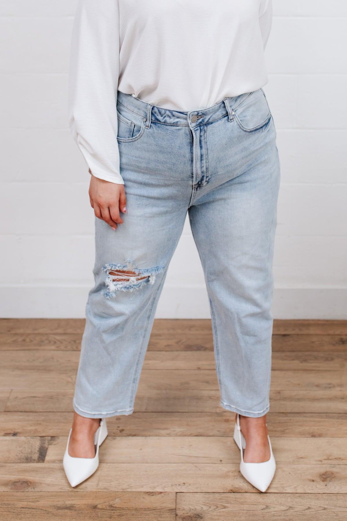 Light blue high-rise jeans with a distressed rip on one thigh, styled with a loose white top and white pointed-toe heels on wooden flooring.
