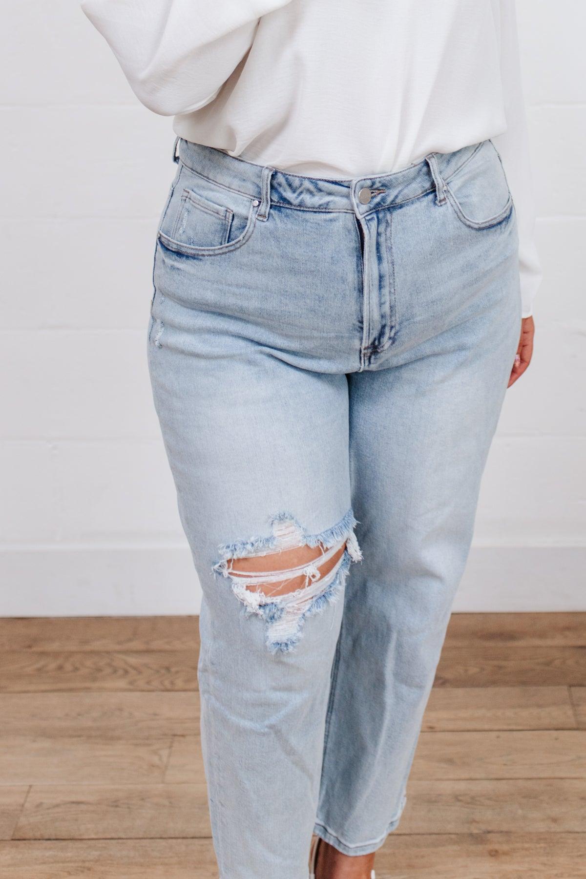 Light blue distressed jeans featuring a large ripped detail on the thigh, styled with a white top and standing on a wooden floor.