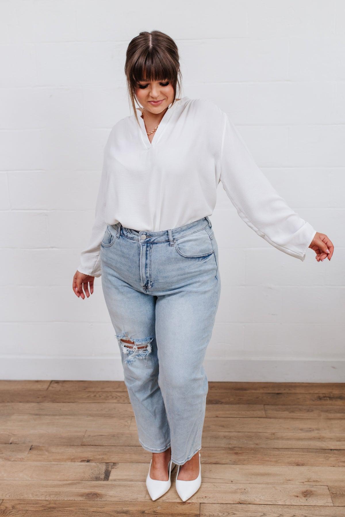 Woman wearing light blue distressed jeans with ripped detail on one thigh paired with a loose white blouse and white pointed heels standing indoors