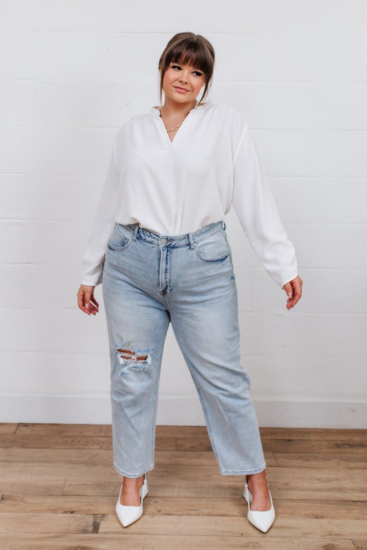 Woman wearing light blue distressed jeans with ripped details on one thigh paired with a loose white blouse and white pointed heels indoors against a white wall