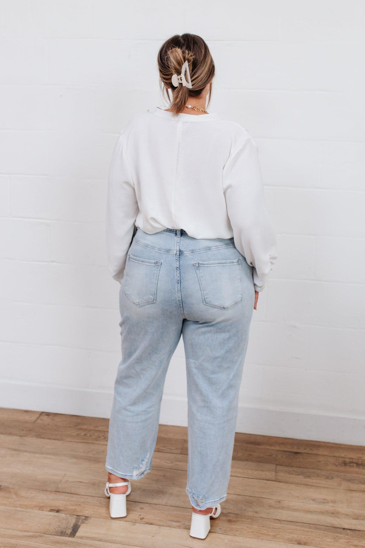 Woman wearing light blue high-rise jeans styled with a loose white long-sleeve top and white heeled sandals, standing indoors against a white wall.