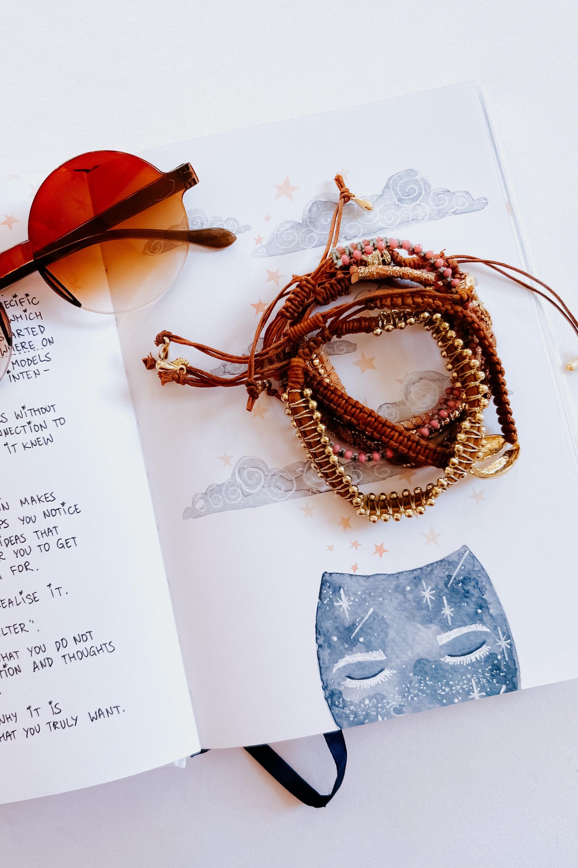 Brown woven beaded stackable bracelet set with gold accents styled on an open illustrated notebook with sunglasses nearby