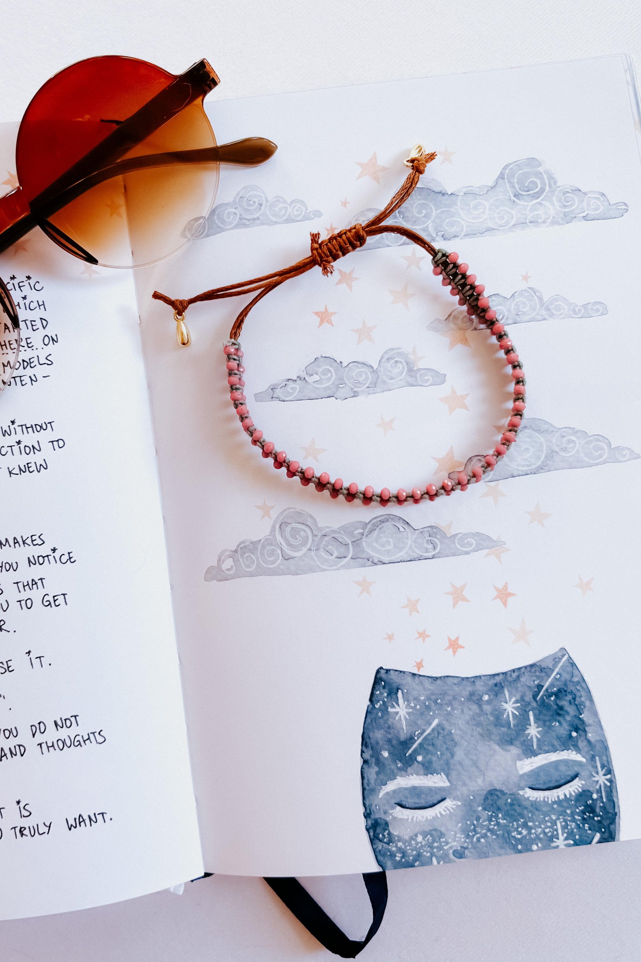 Brown and pink beaded woven bracelet with gold charm displayed on an illustrated page with clouds and stars nearby