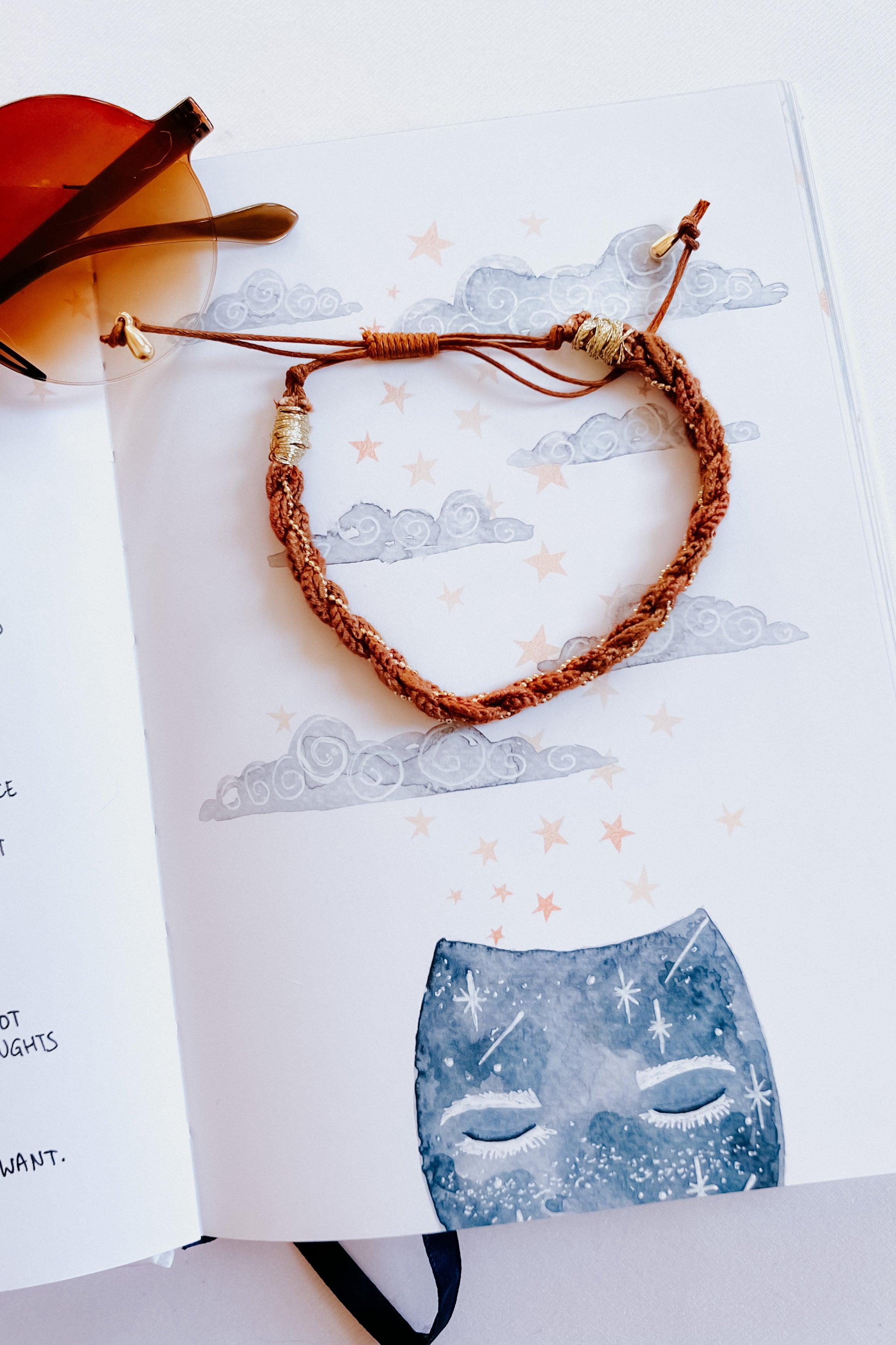 Brown woven bracelet with gold accents styled on an open illustrated journal featuring stars and clouds artwork