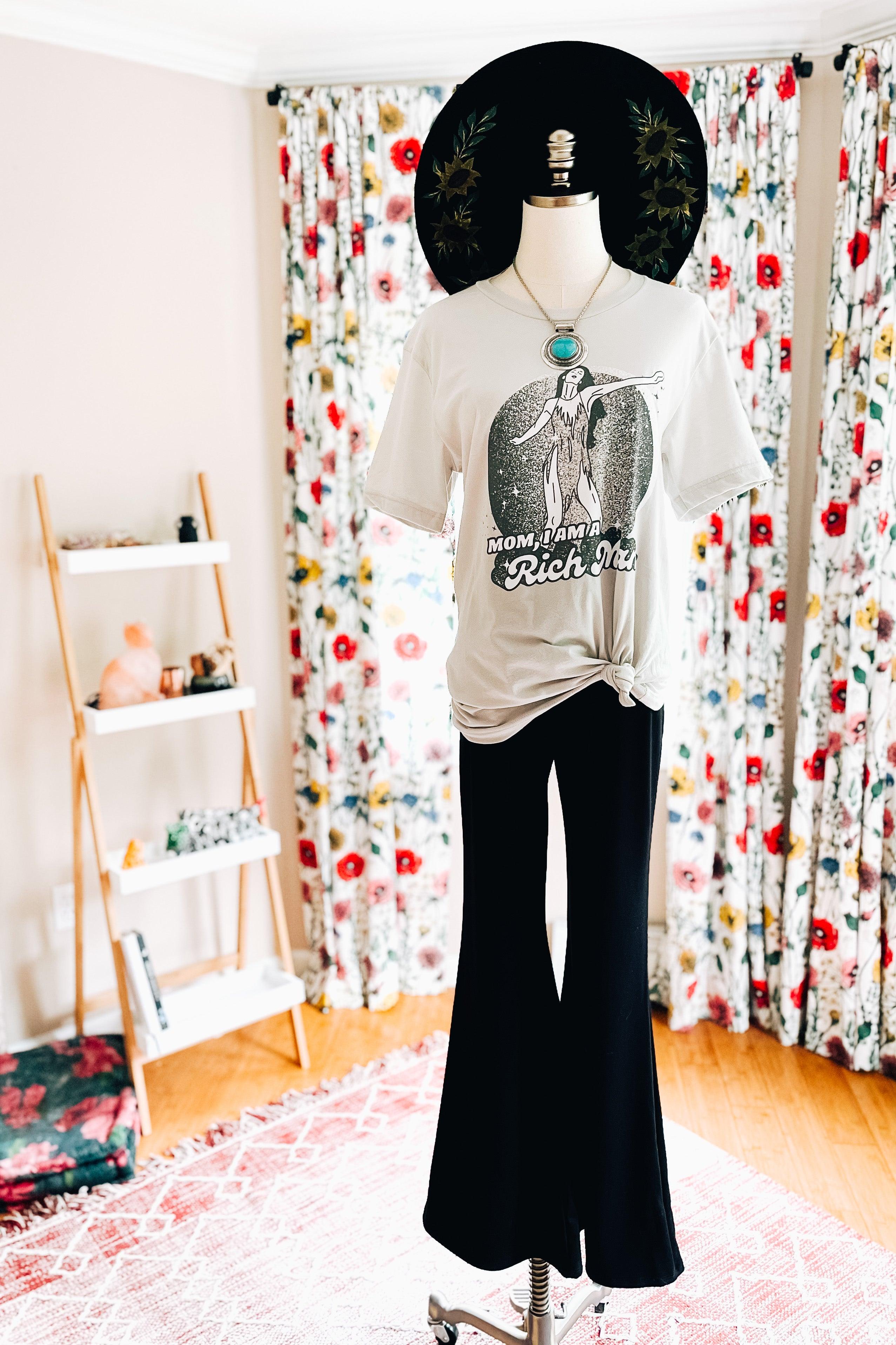 Mannequin wearing black flared pants with knotted graphic tee, turquoise necklace, and wide-brimmed black hat by floral curtains