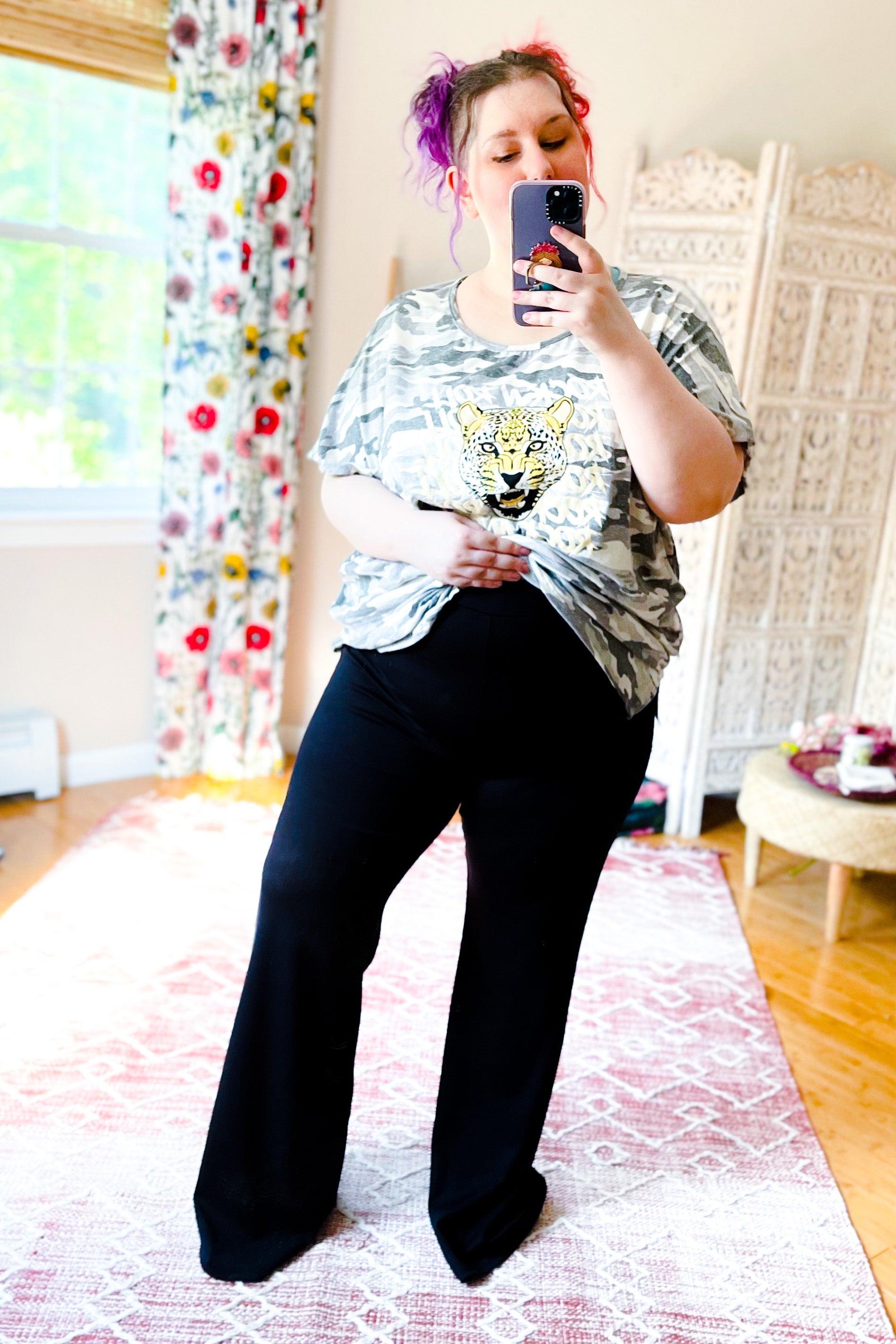 Woman with pink and purple hair wearing black flare pants and animal print top taking mirror selfie in bright room