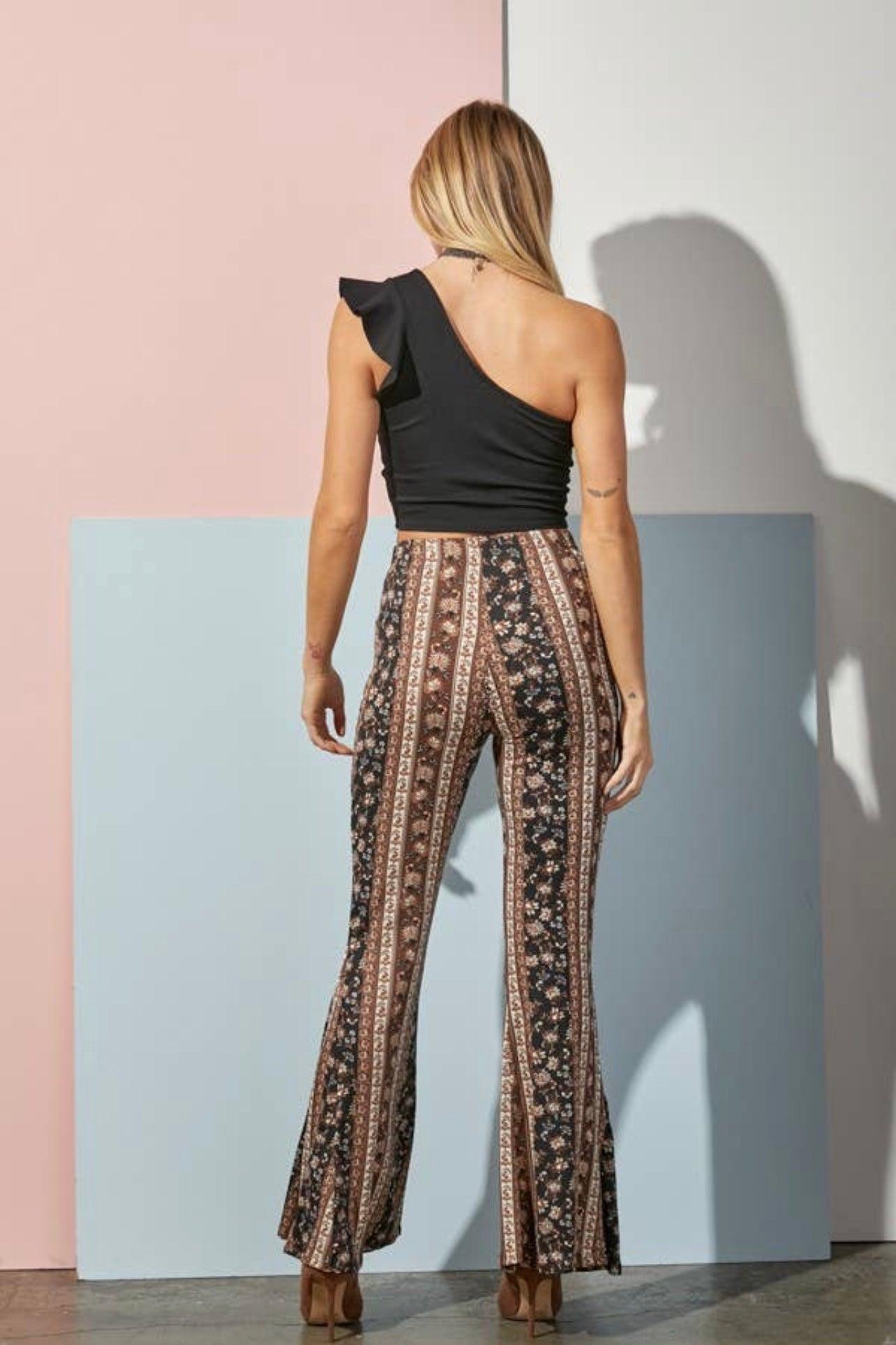 Woman wearing navy and brown striped paisley pants with high waist and black one-shoulder ruffle top standing against pastel wall