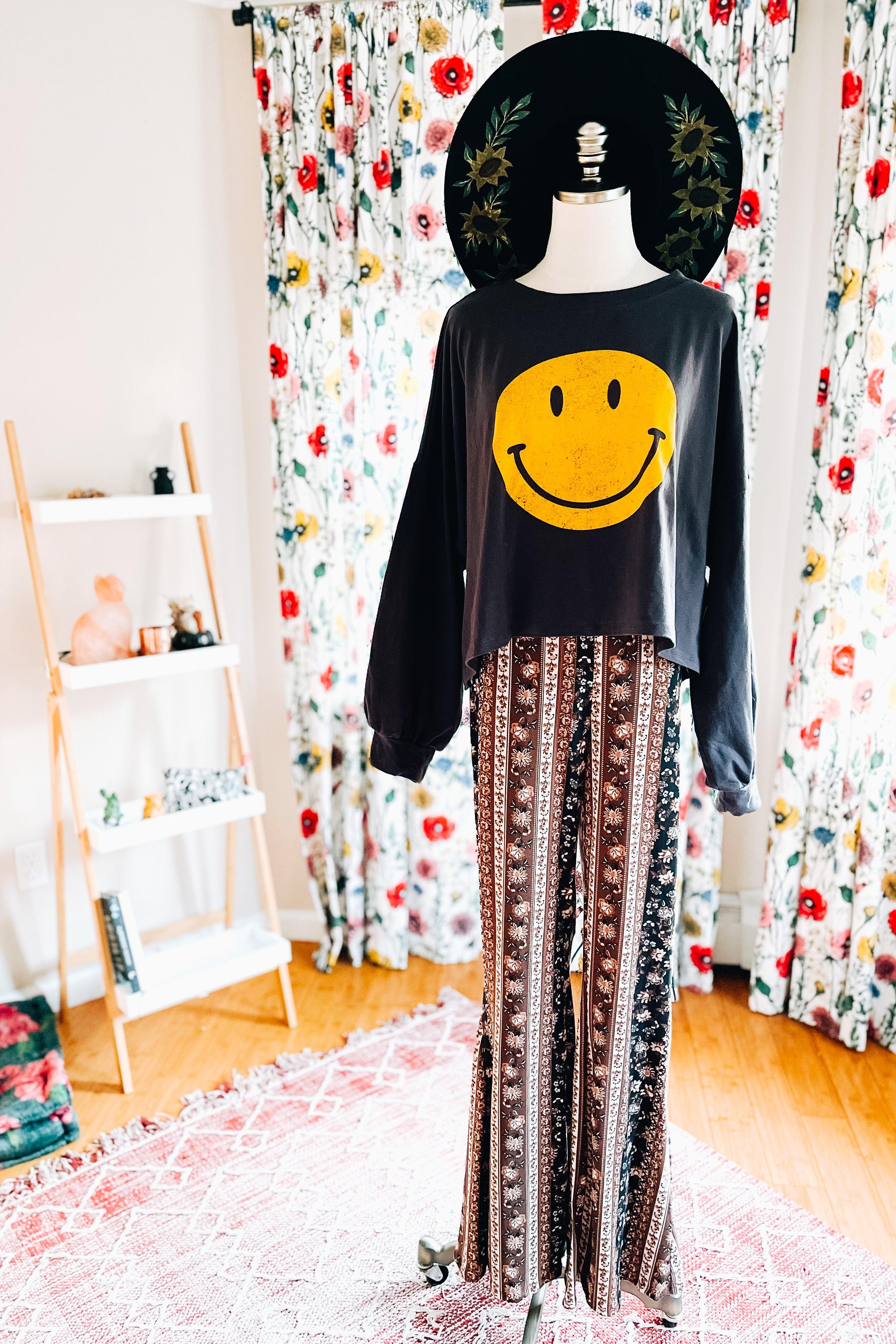 Mannequin dressed in navy and brown paisley bell bottoms with a black smiley face long sleeve top and wide-brim hat indoors