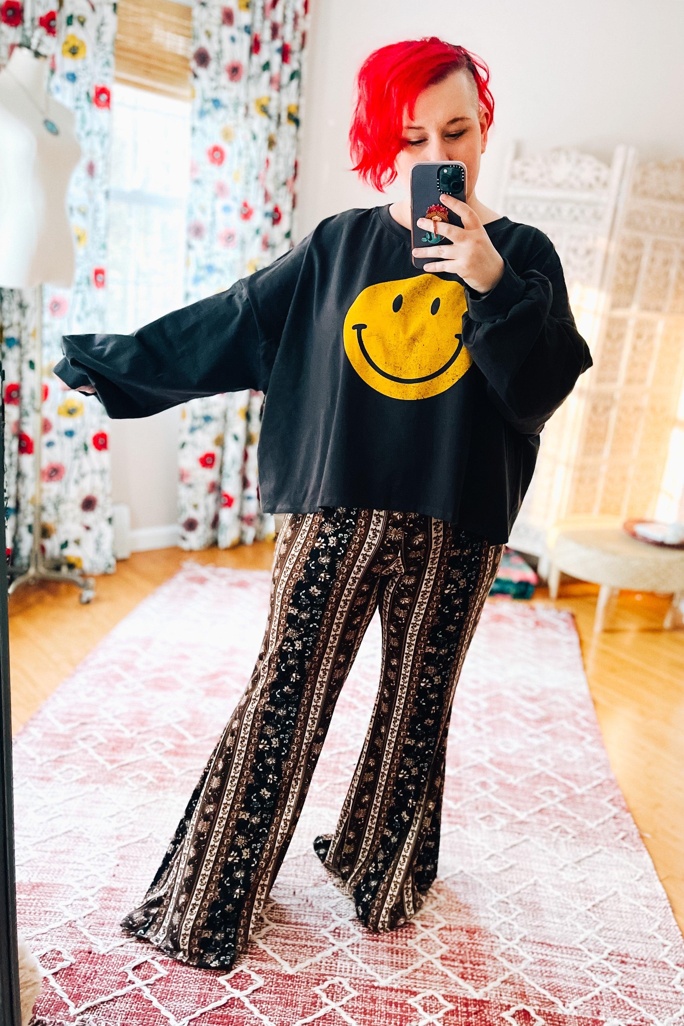 Woman with bright red hair wearing navy and brown paisley-striped bell bottoms and a black smiley face sweatshirt indoors