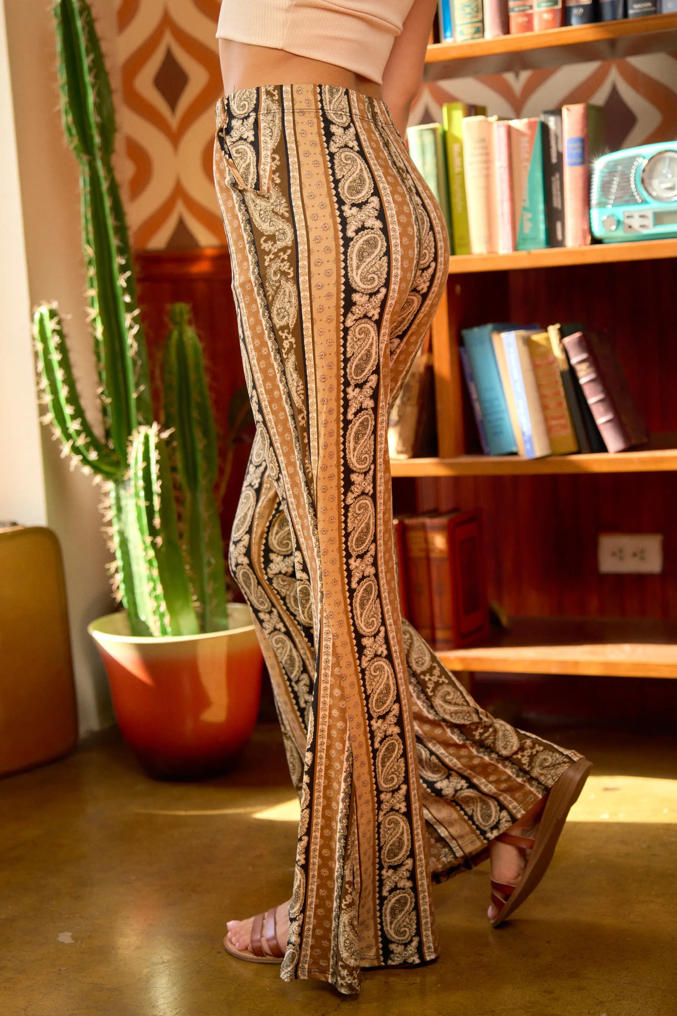 Person wearing rust paisley print flared pants and cropped blush top in a room with cactus and retro bookcase, bold eclectic style