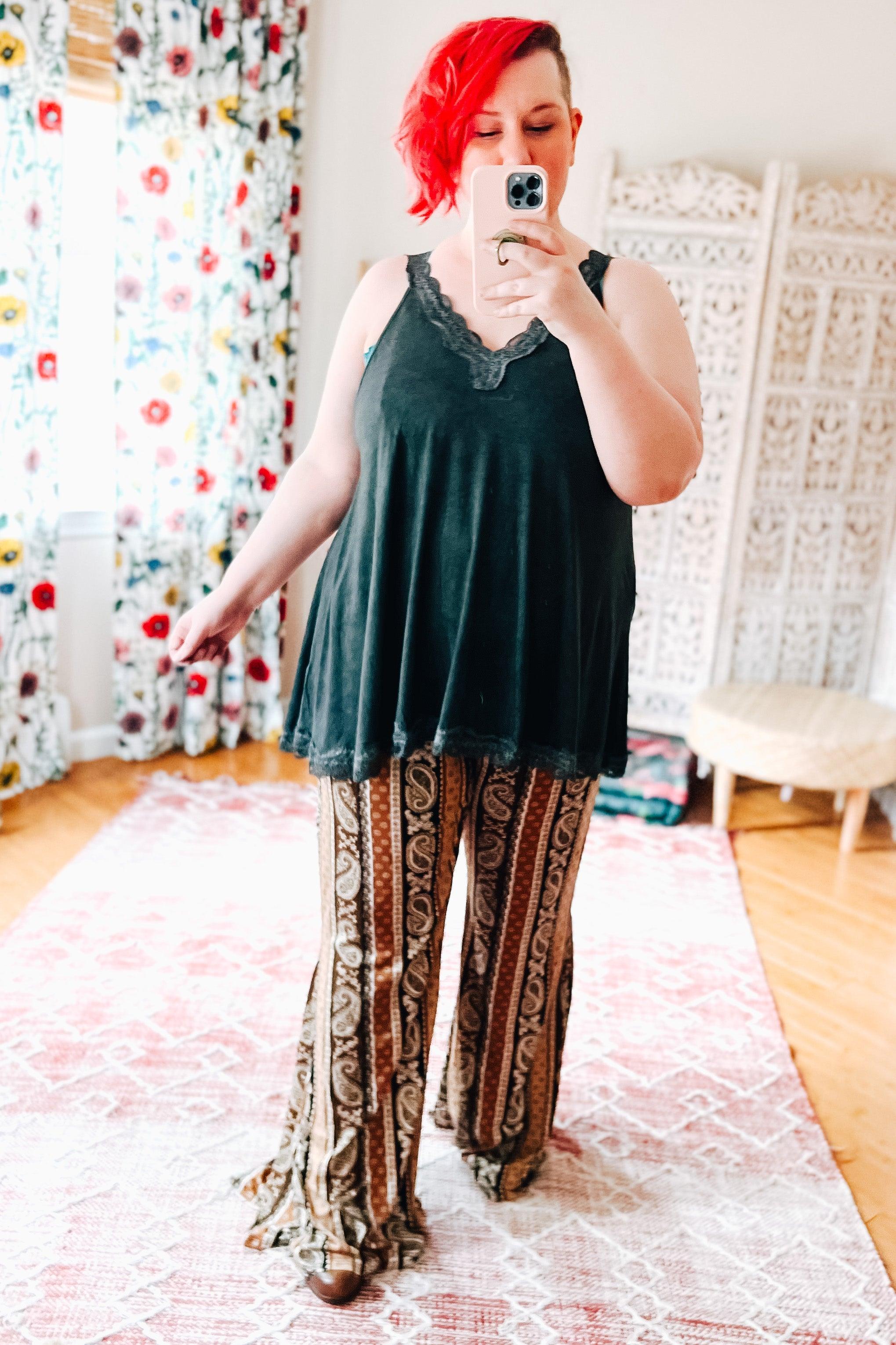 Woman with bright red hair wearing black lace-trimmed tank and rust paisley print flared pants in cozy room setting