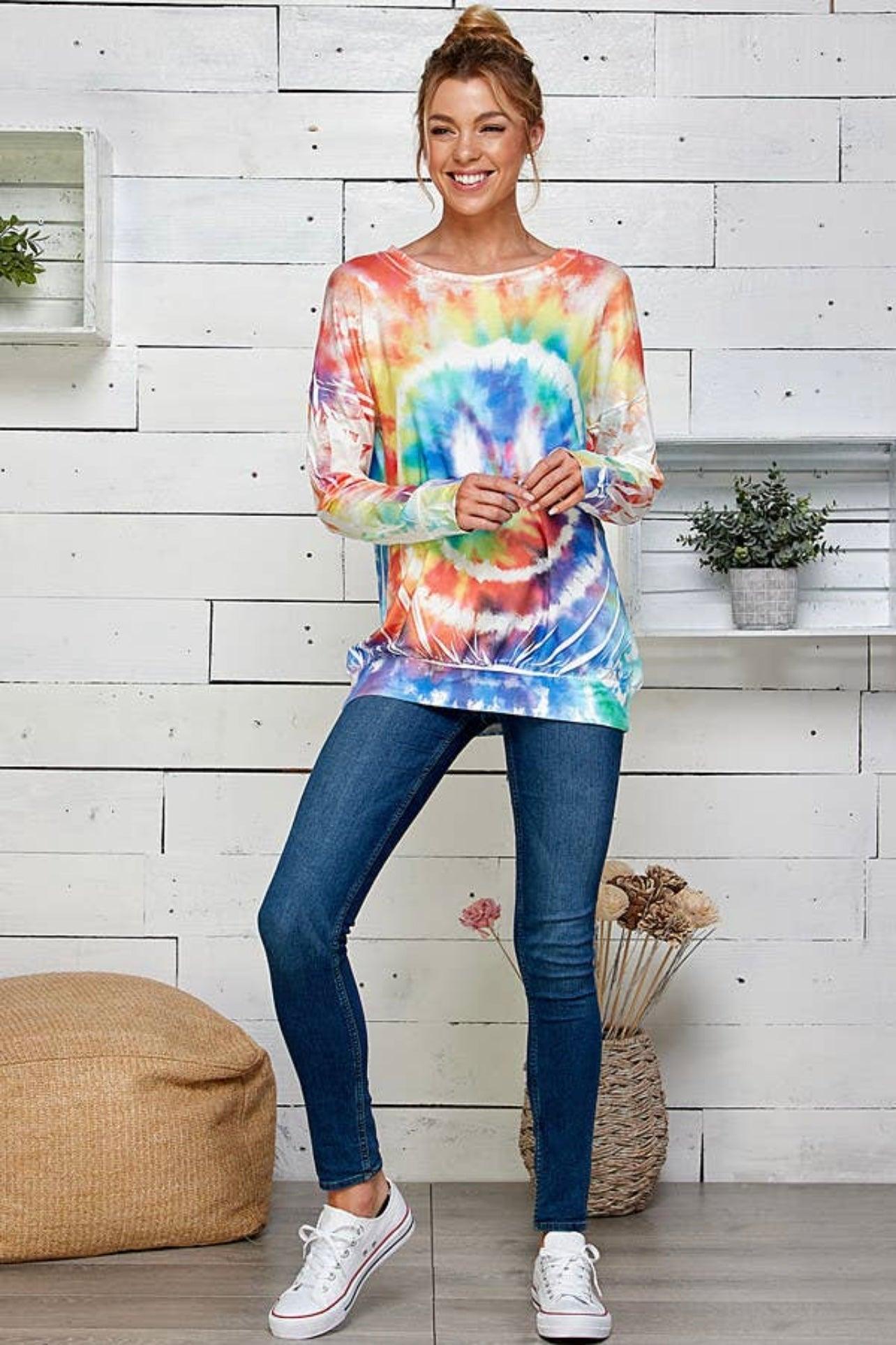 Smiling woman wearing a tie dye rainbow pullover styled with skinny jeans and white sneakers in a bright room