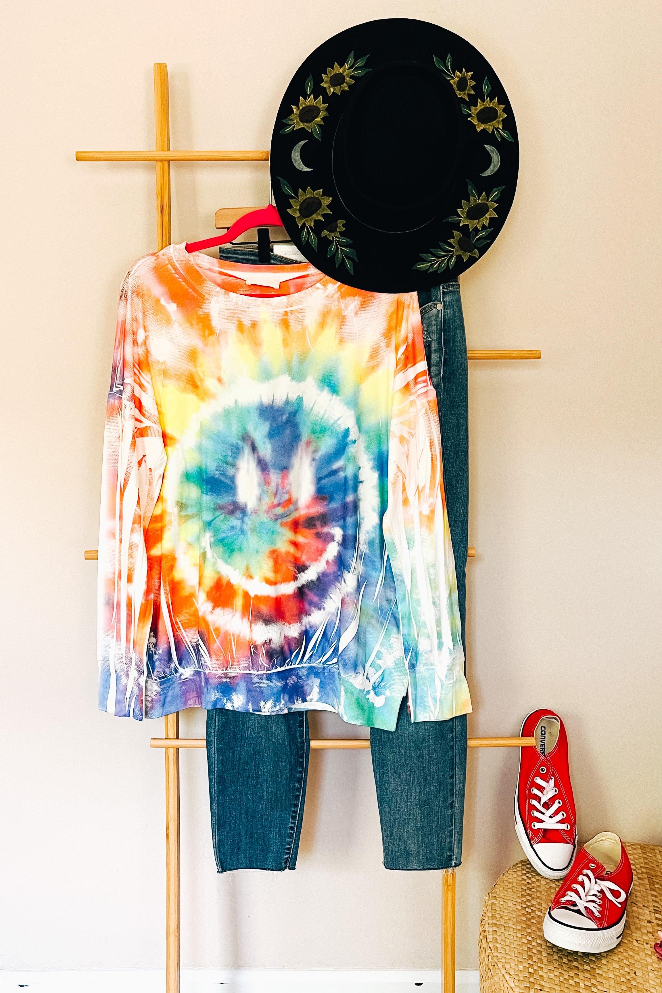 Tie dye rainbow pullover with smiley design styled with denim and sunflower detail black hat indoors