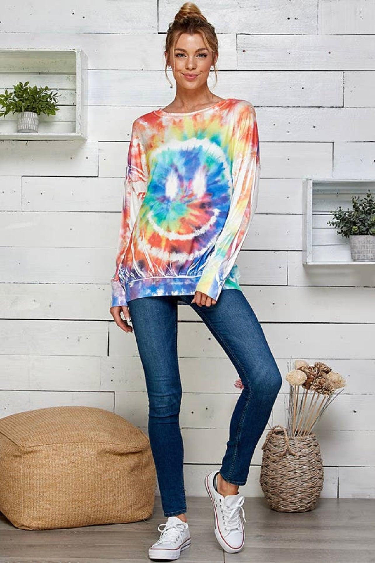 Woman wearing a tie dye rainbow pullover with smiley design paired with slim jeans and white sneakers in cozy setting