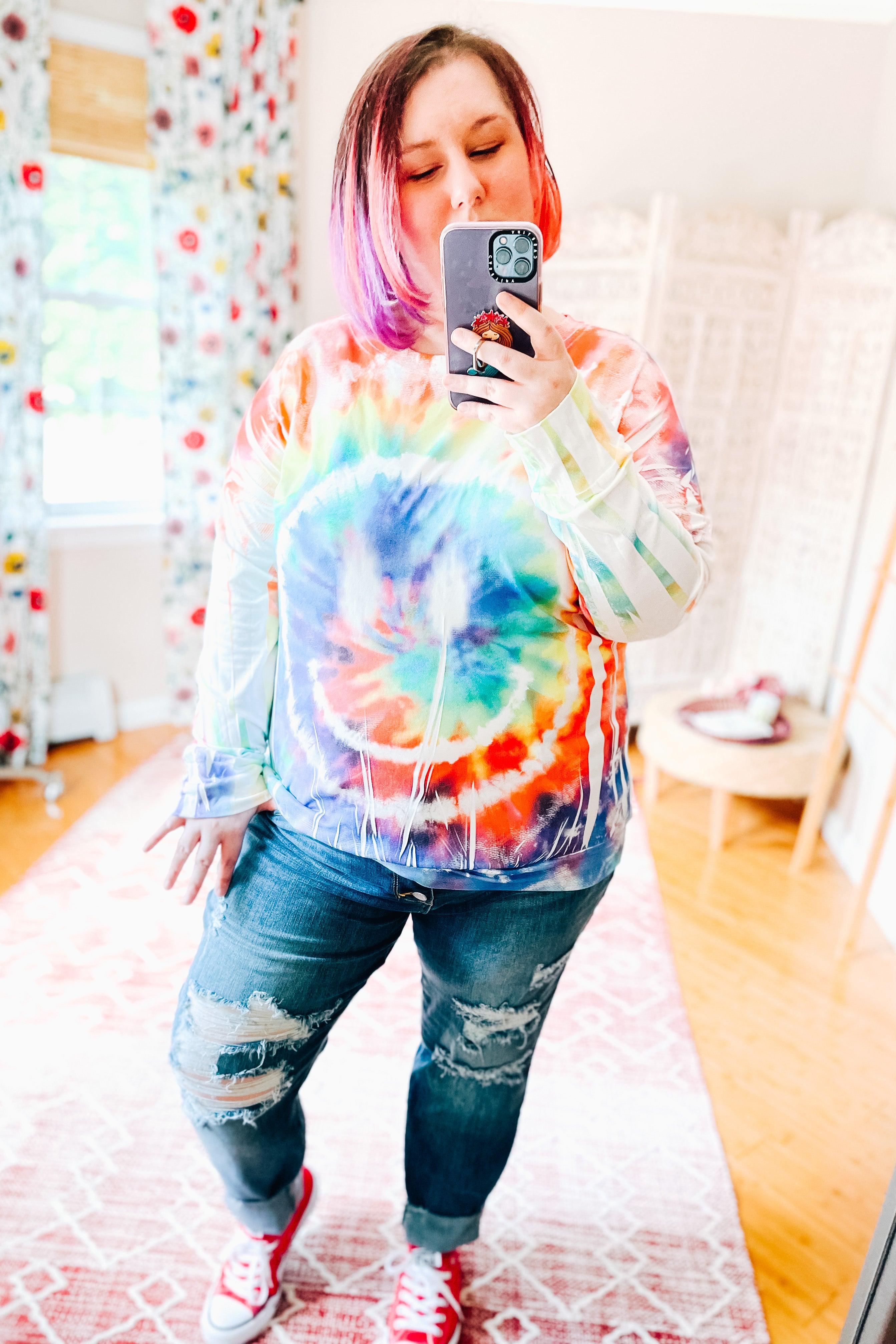 Plus size woman wearing a colorful tie dye rainbow pullover with a circular pattern and distressed jeans indoors