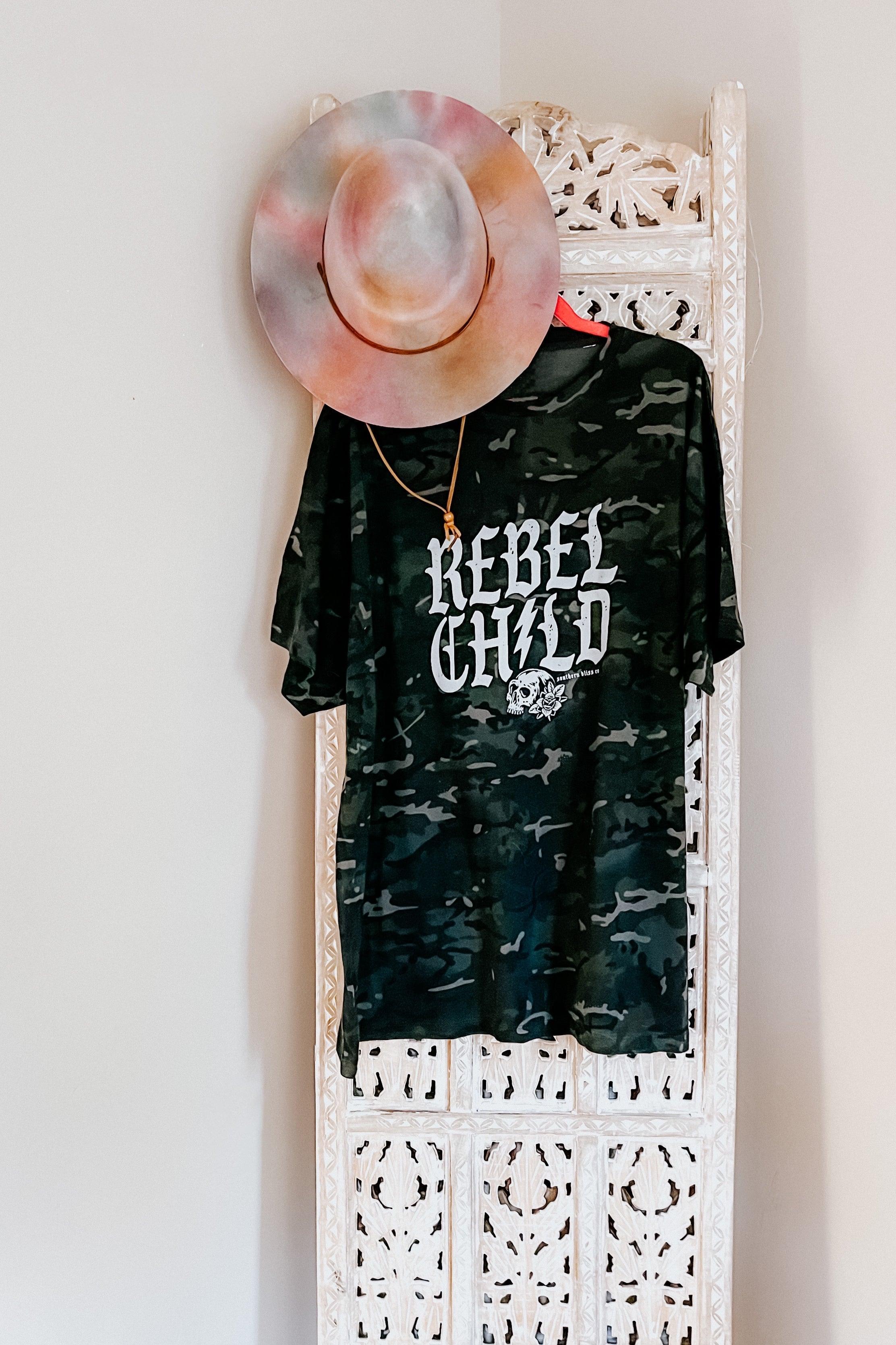 Camo green oversized tee with bold white “Rebel Child” skull print hanging on carved white wooden screen with hat and necklace