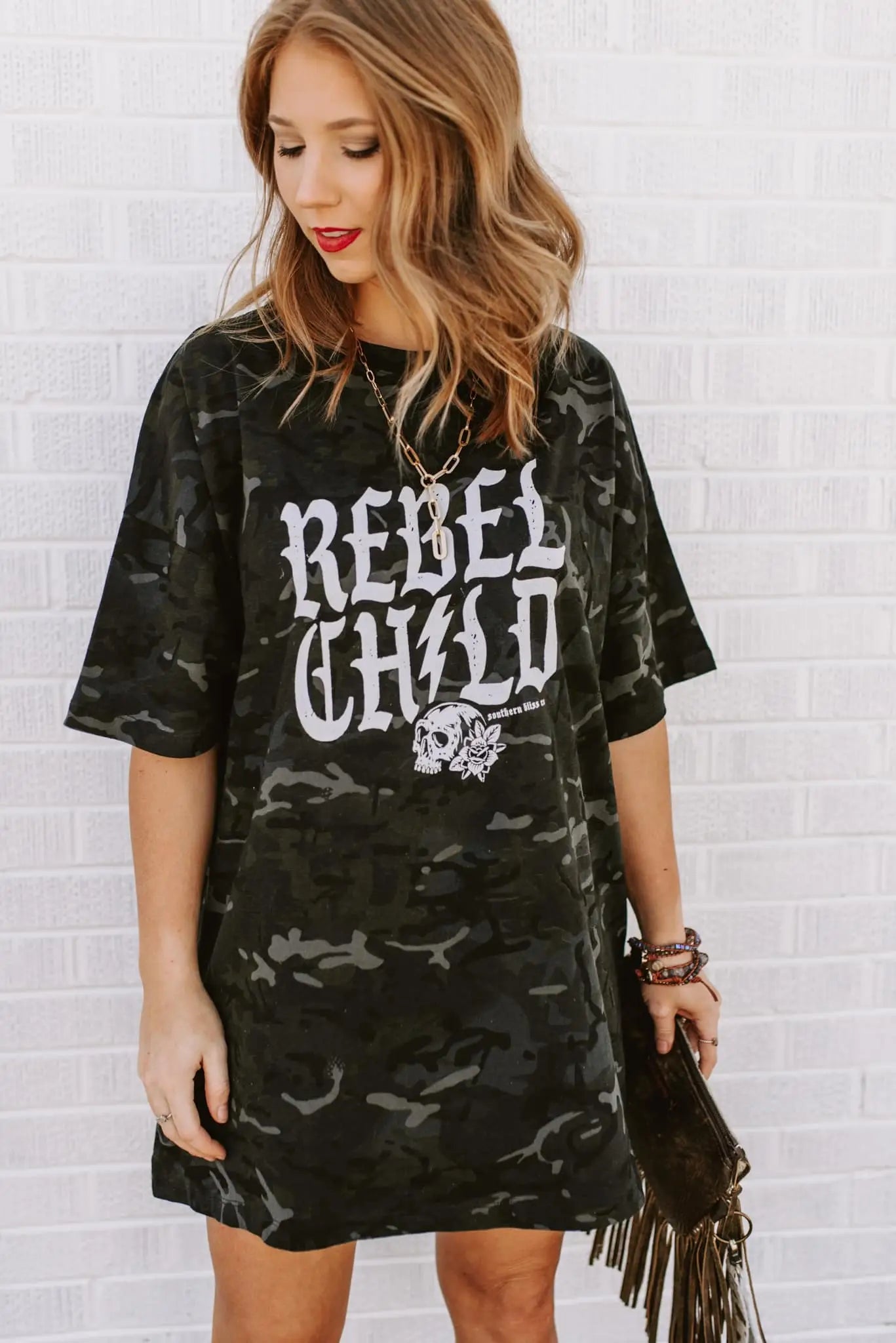 Woman wearing oversized camo green tee with bold skull and “Rebel Child” text styled with necklace and wristbands