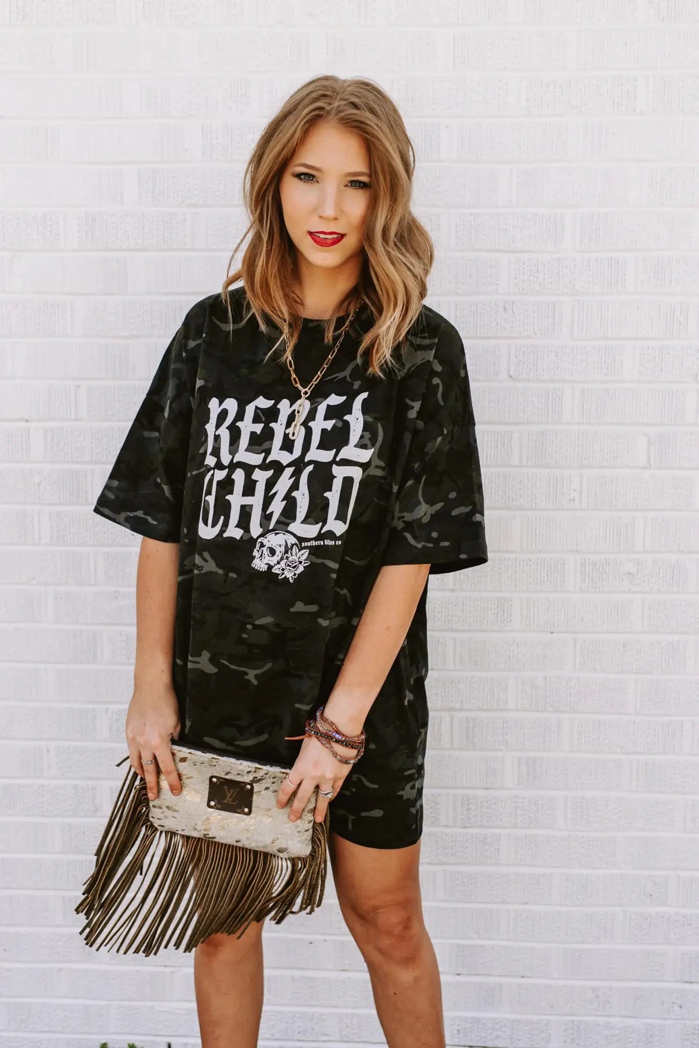 Woman wearing camo green oversized tee with bold "Rebel Child" lettering styled with fringe bag and layered bracelets