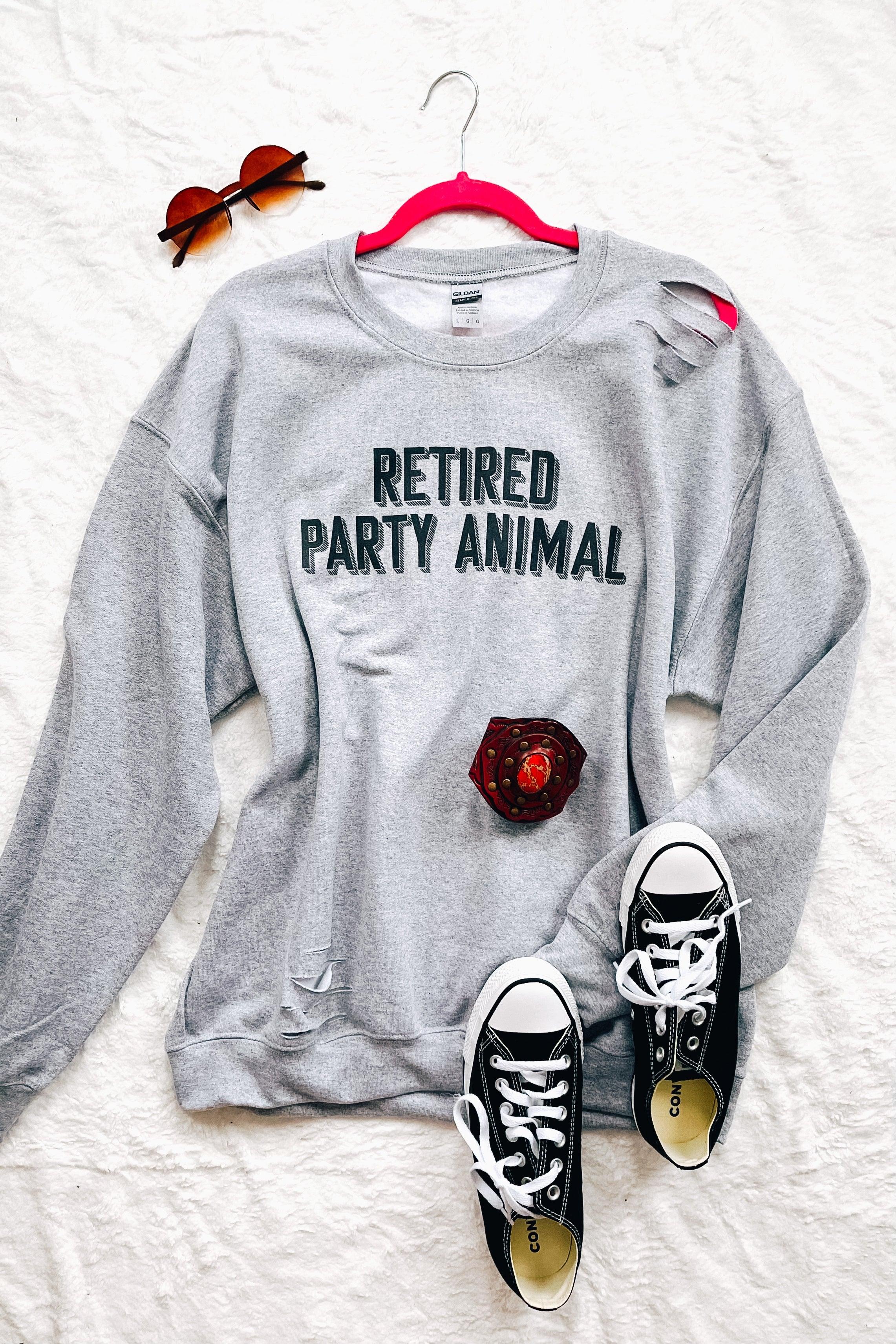 Grey distressed pullover with bold "Retired Party Animal" text styled with black shoes and sunglasses on white background