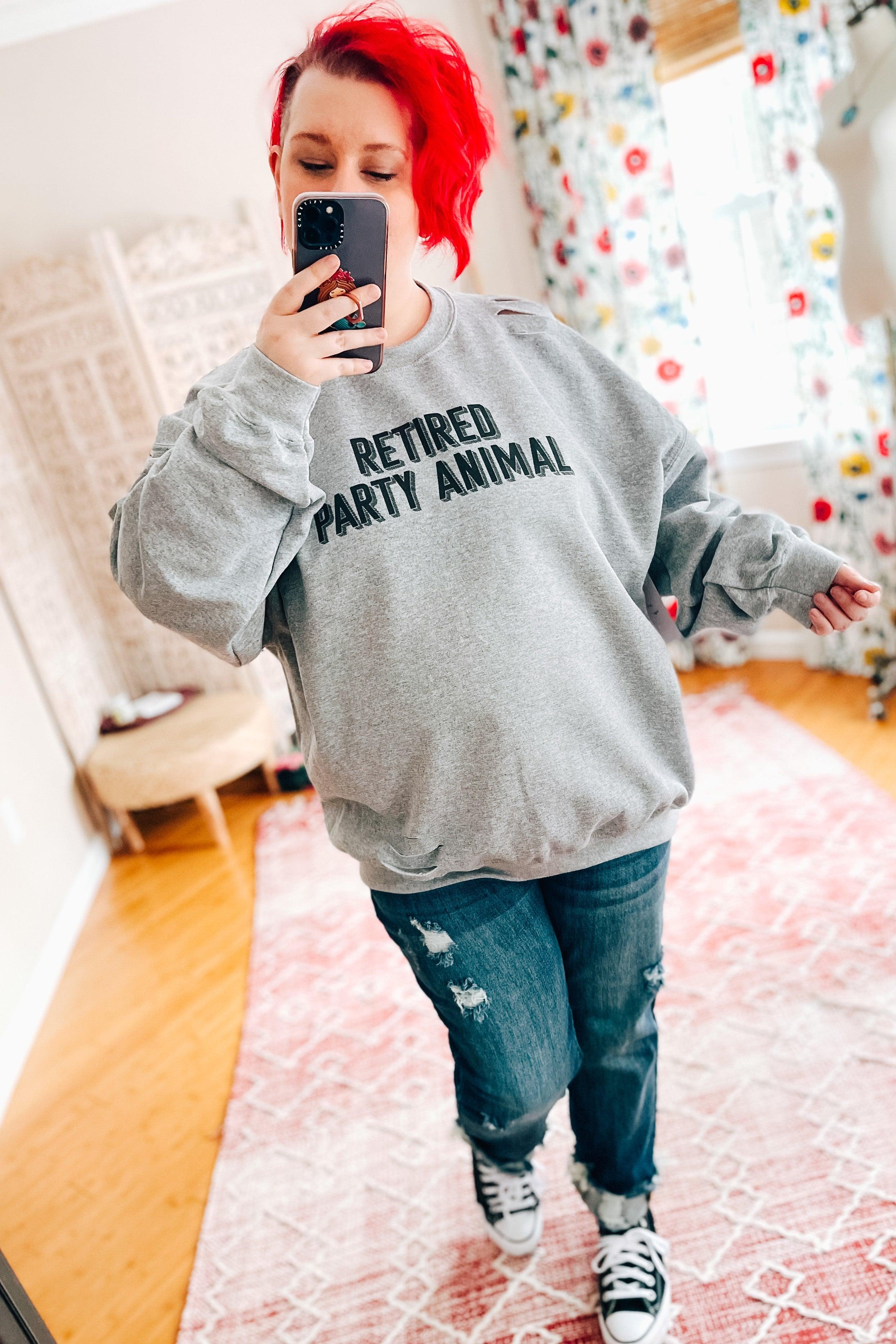 Woman with bright red hair wearing grey distressed pullover with bold text taking mirror selfie indoors on pink rug