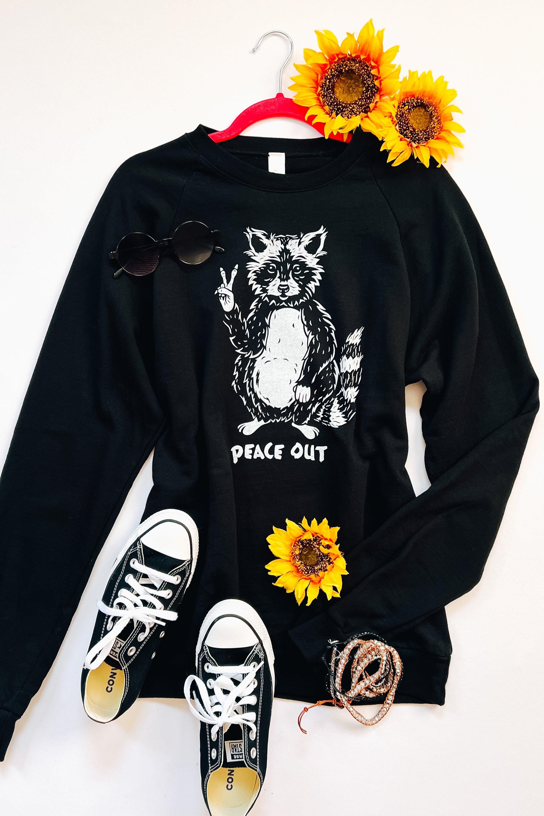 Black organic cotton sweatshirt with raccoon graphic making peace sign and “peace out” text styled with sunglasses and sneakers