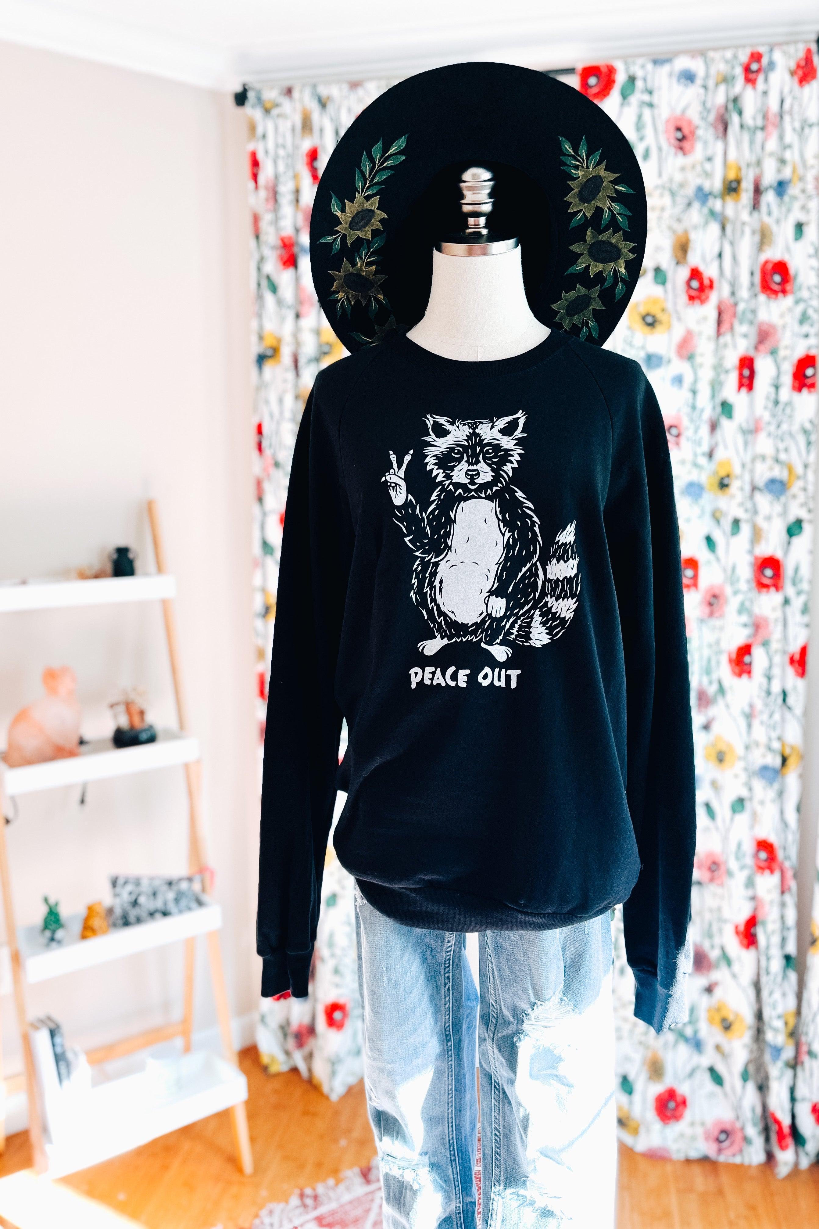 Black organic cotton sweatshirt with raccoon flashing peace sign and “peace out” text styled on mannequin with floral hat and jeans
