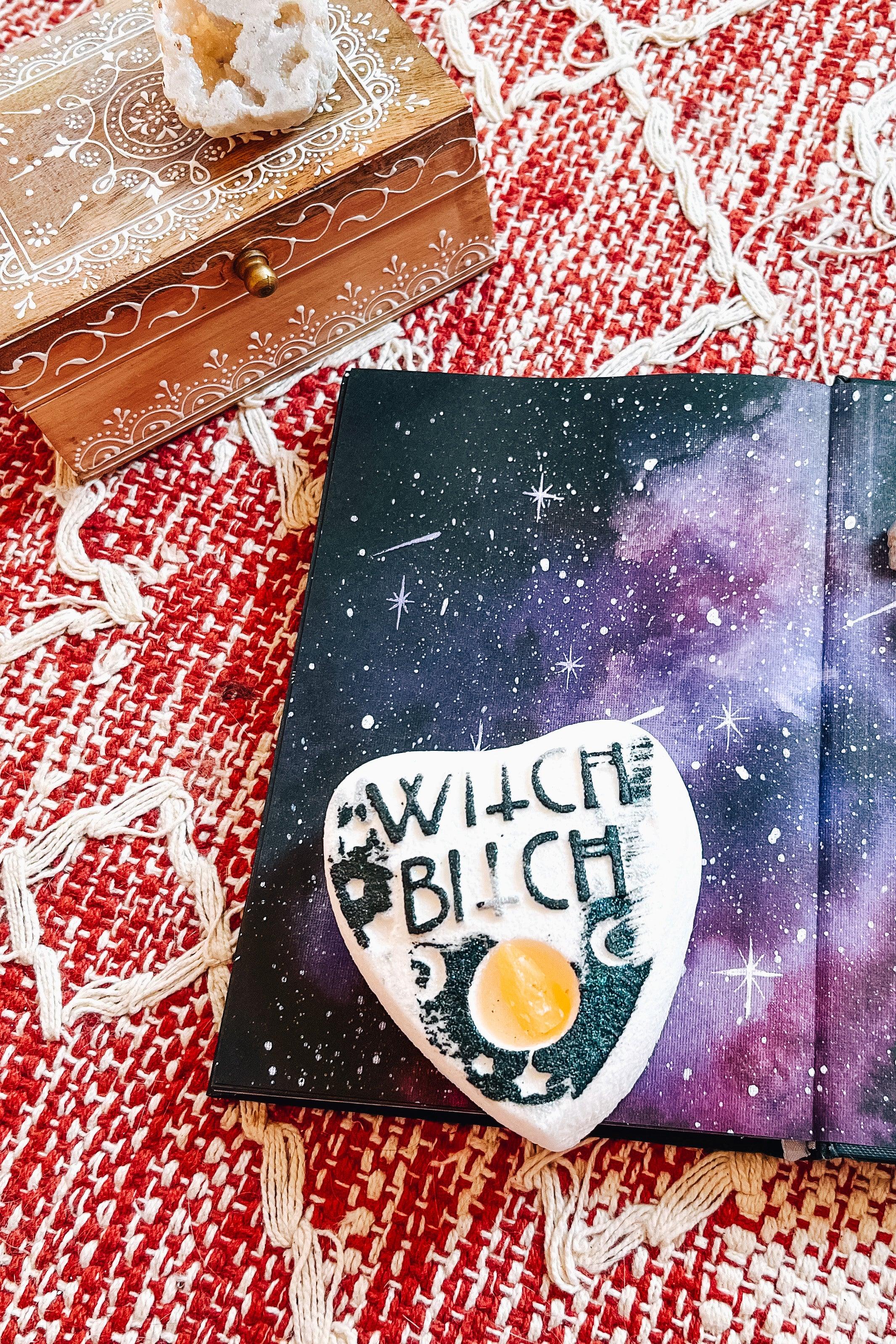 Planchette-shaped white bath bomb with black lettering saying “witch bitch” and citrine crystal center set on galaxy-patterned book atop woven red textile