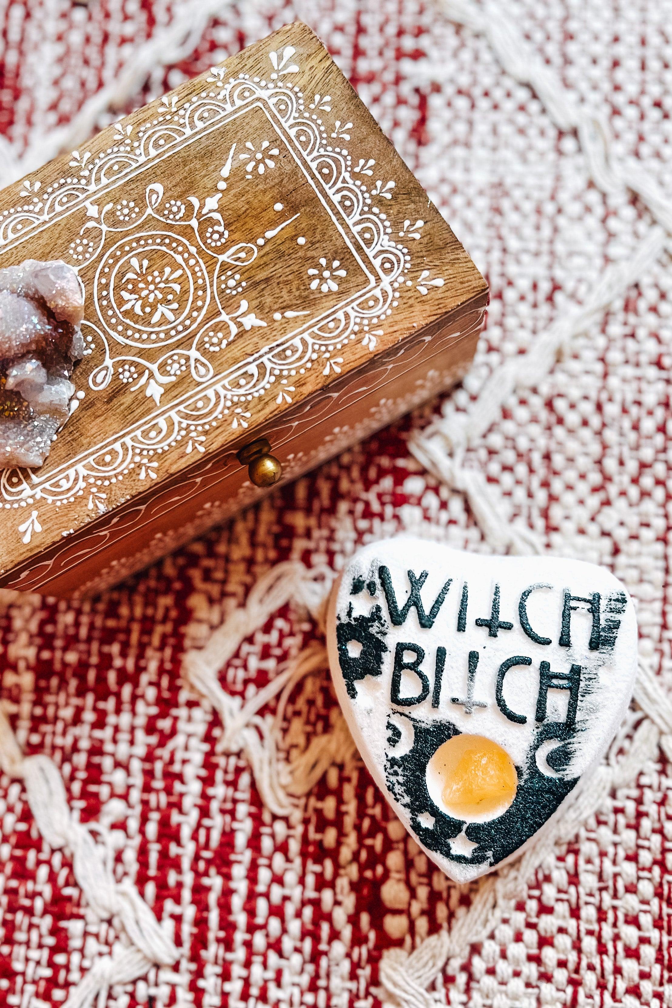 White planchette-shaped bath bomb with black lettering that says “witch bitch” and a citrine crystal beside a carved wooden box on a textured fabric