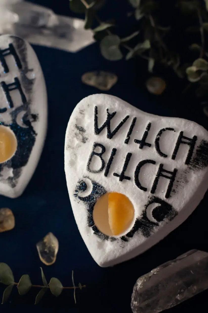 White planchette-shaped bath bomb with black lettering that says “witch bitch” and crescent moon symbols on dark surface with crystals and greenery