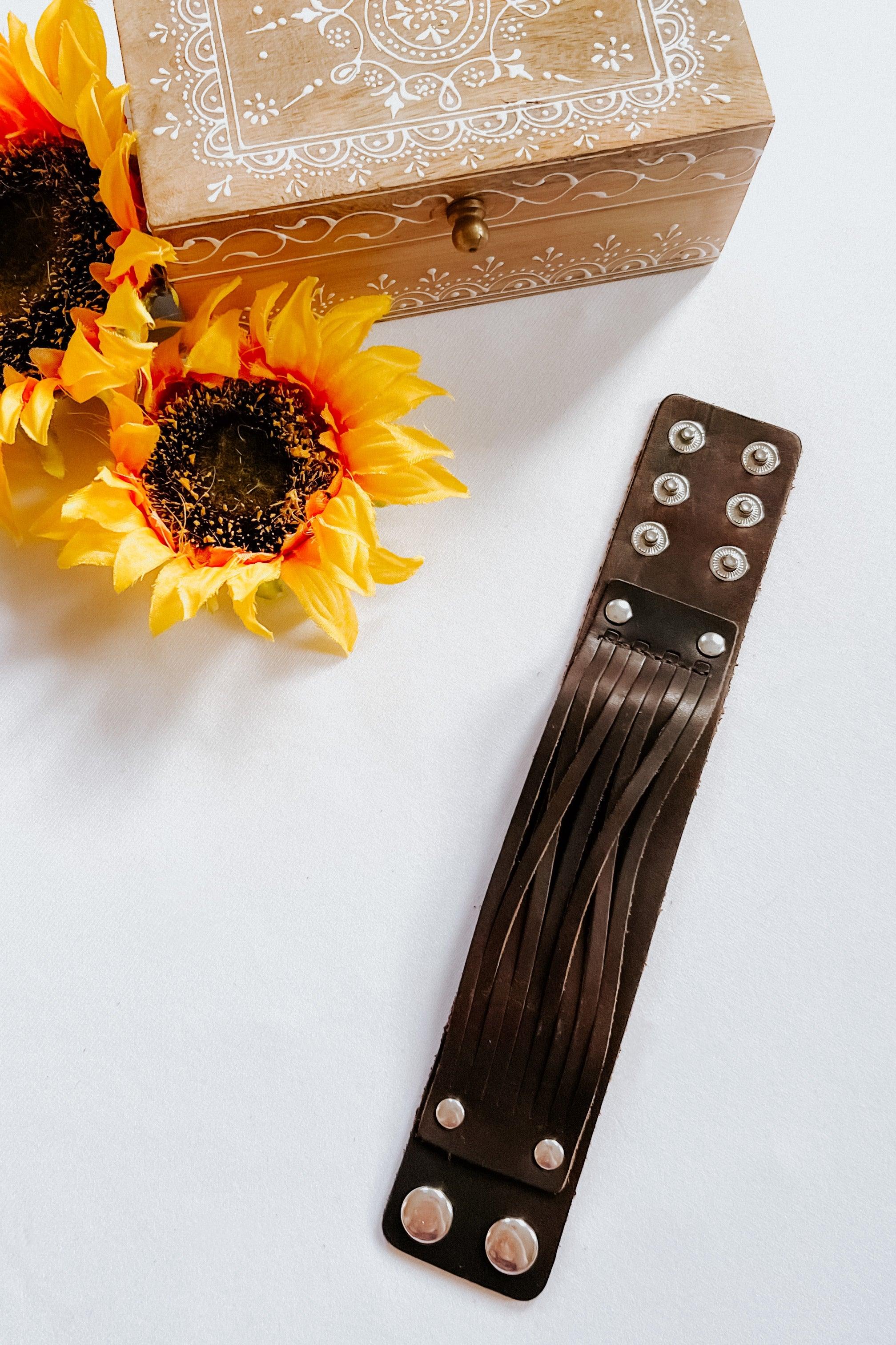 Brown leather cuff bracelet with multiple straps and metal snaps displayed beside sunflowers and wooden box