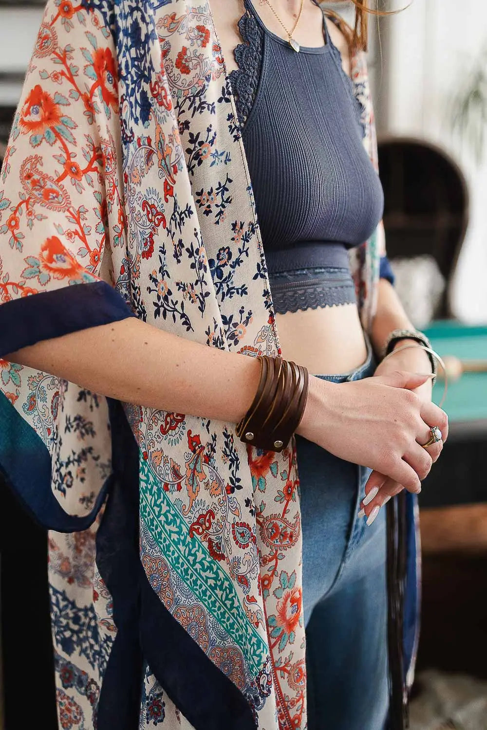 Woman wearing layered floral kimono and navy crop top with brown leather cuff bracelet and casual jeans indoors