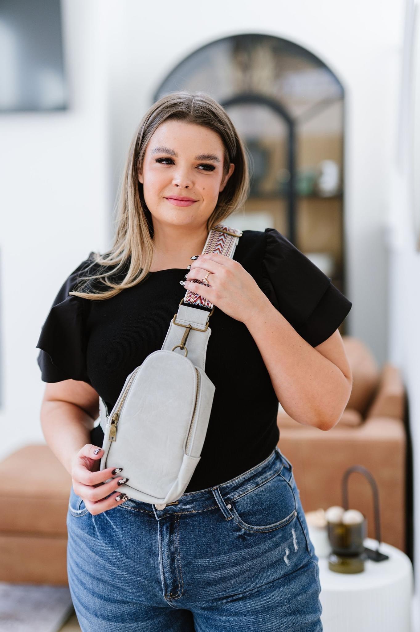 Woman wearing black top and jeans holding a grey vegan leather sling bag with patterned strap indoors
