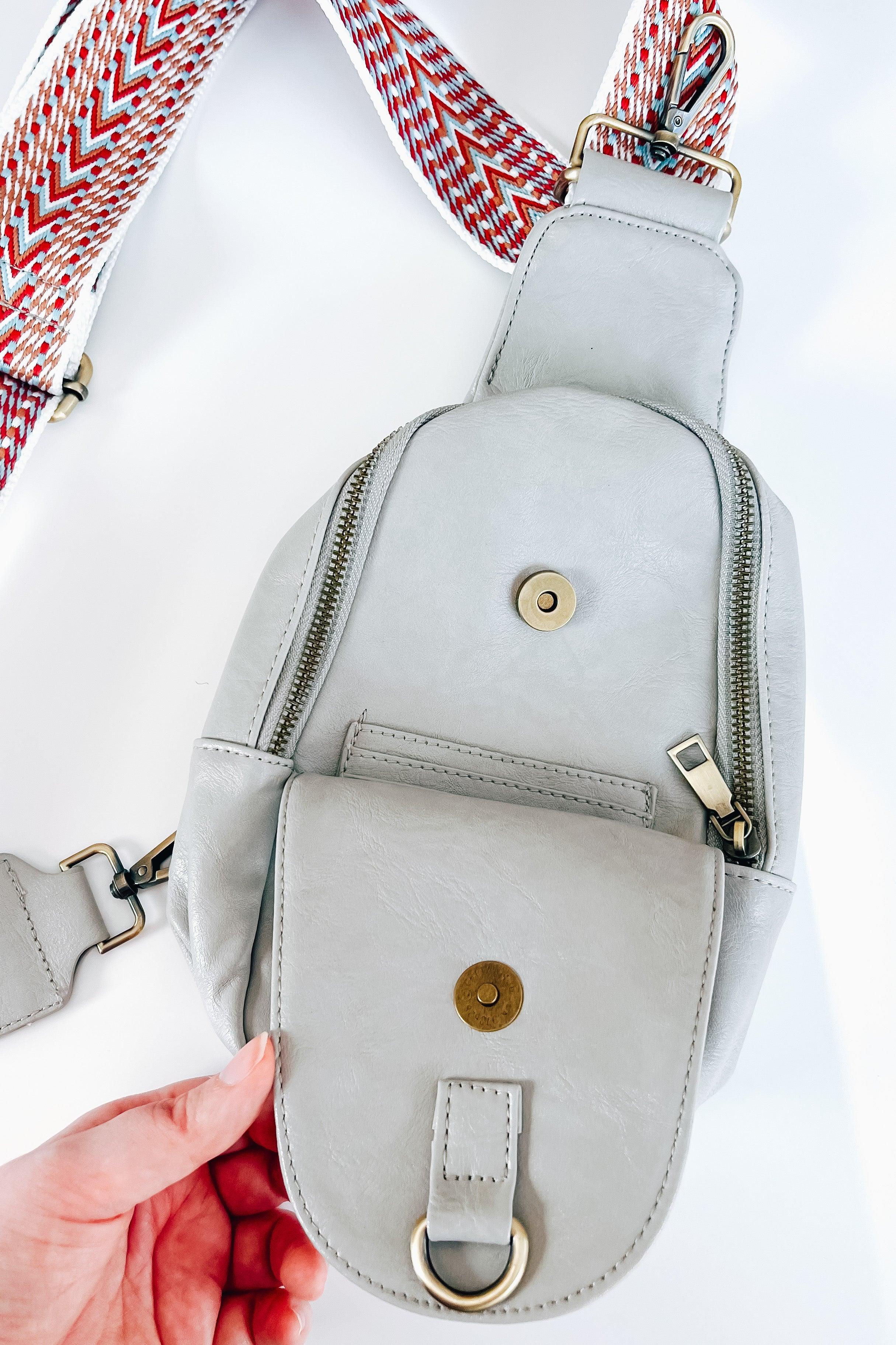 Hand holding gray vegan leather sling bag with front flap pocket and patterned adjustable strap on white background