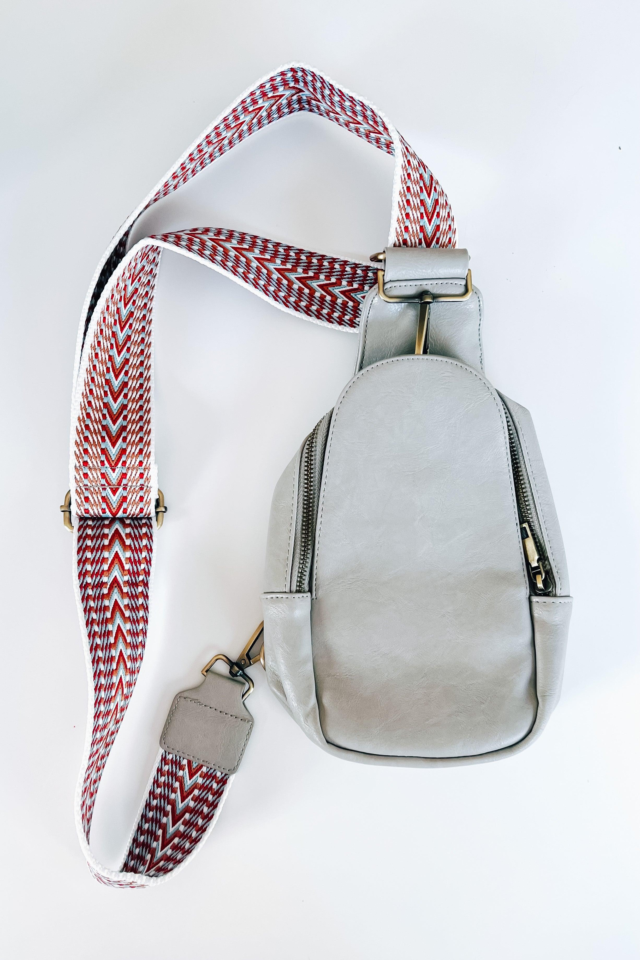 Grey vegan leather sling bag with wide patterned strap in red, white, and black zigzag design on white background