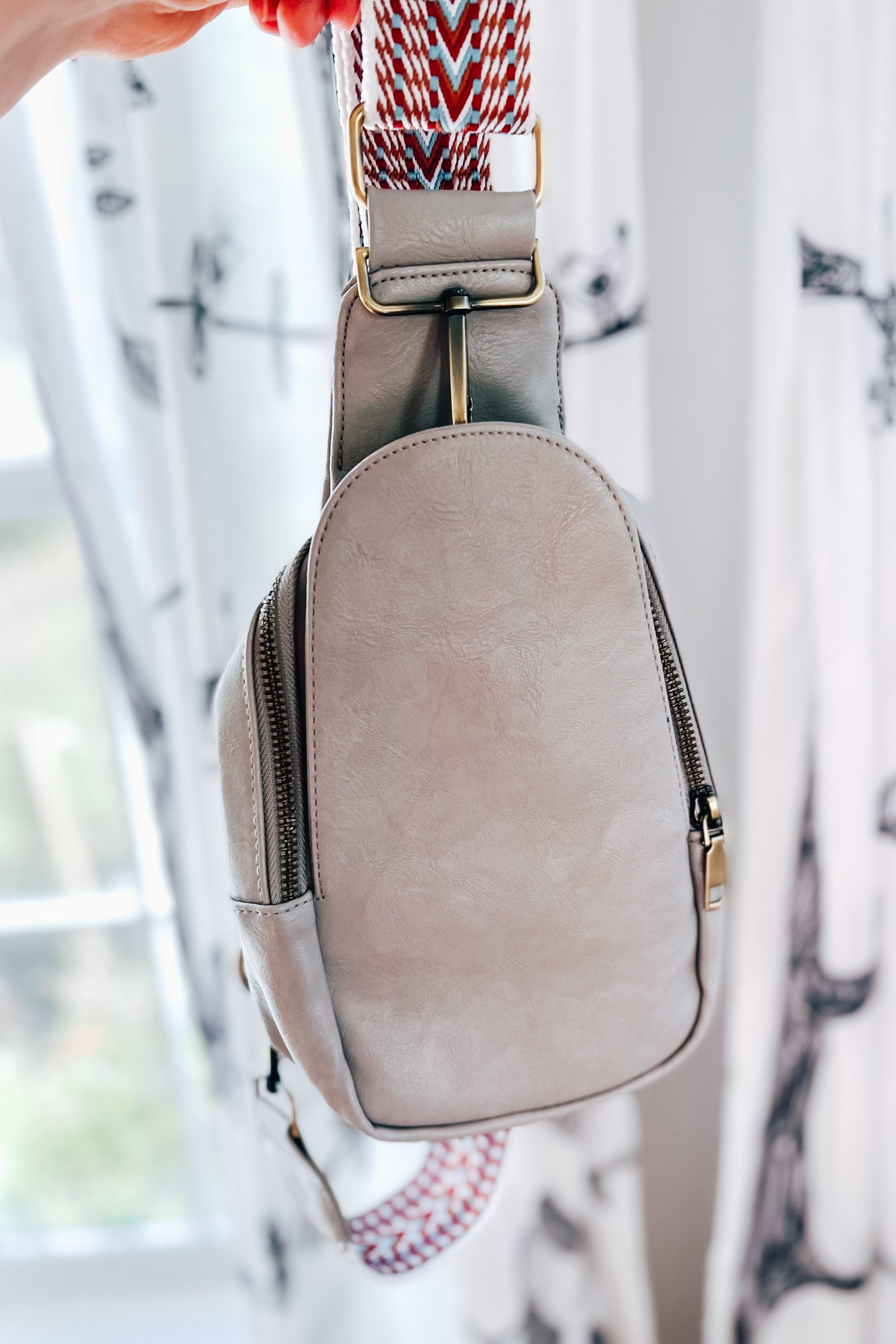 Hand holding a grey vegan leather sling bag with patterned adjustable strap indoors near window light