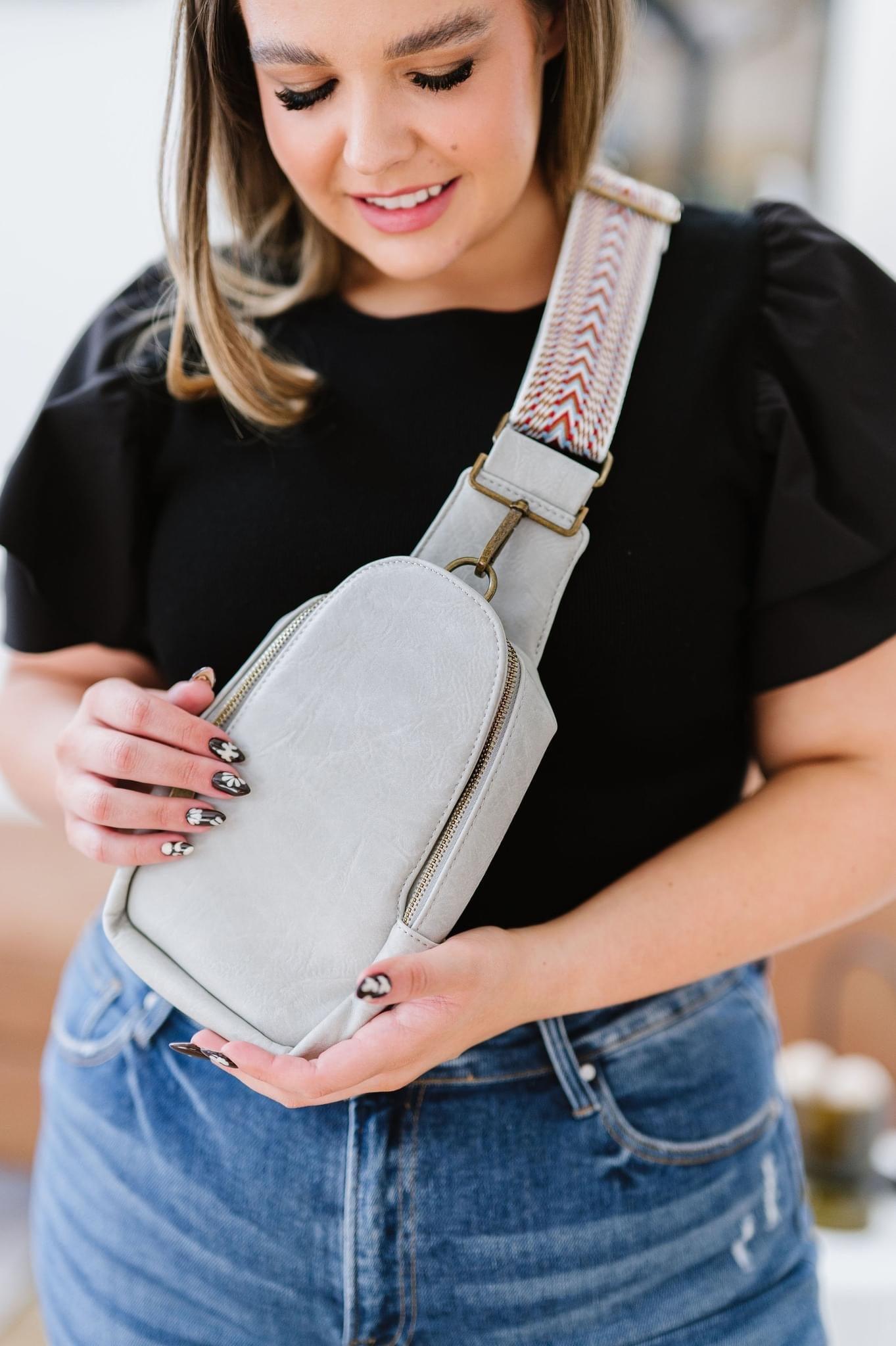 Woman holding grey vegan leather sling bag with patterned strap, wearing black top and blue jeans indoors