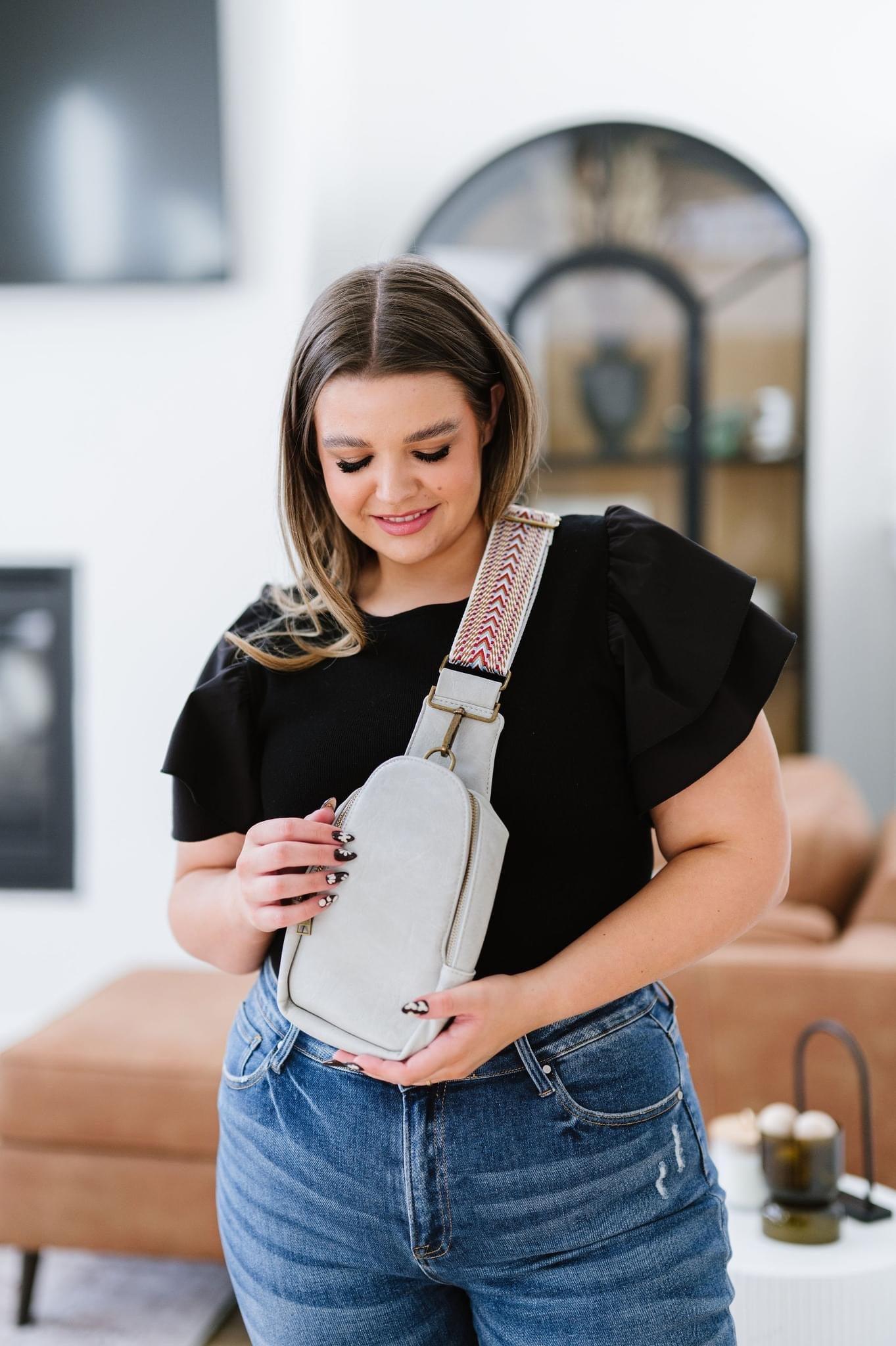 Woman holding grey vegan leather sling bag with patterned strap wearing black top and blue jeans indoors
