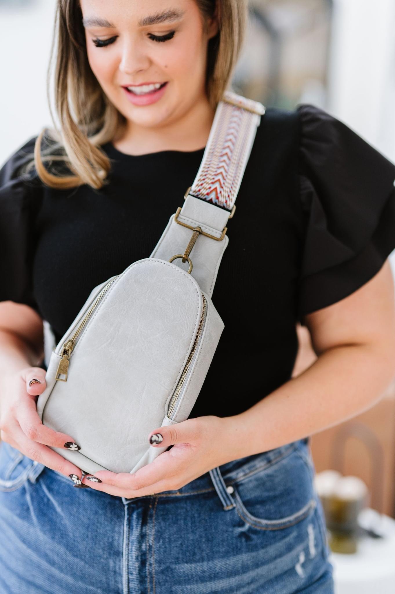 Woman wearing black top and jeans holding gray vegan leather sling bag with patterned strap indoors