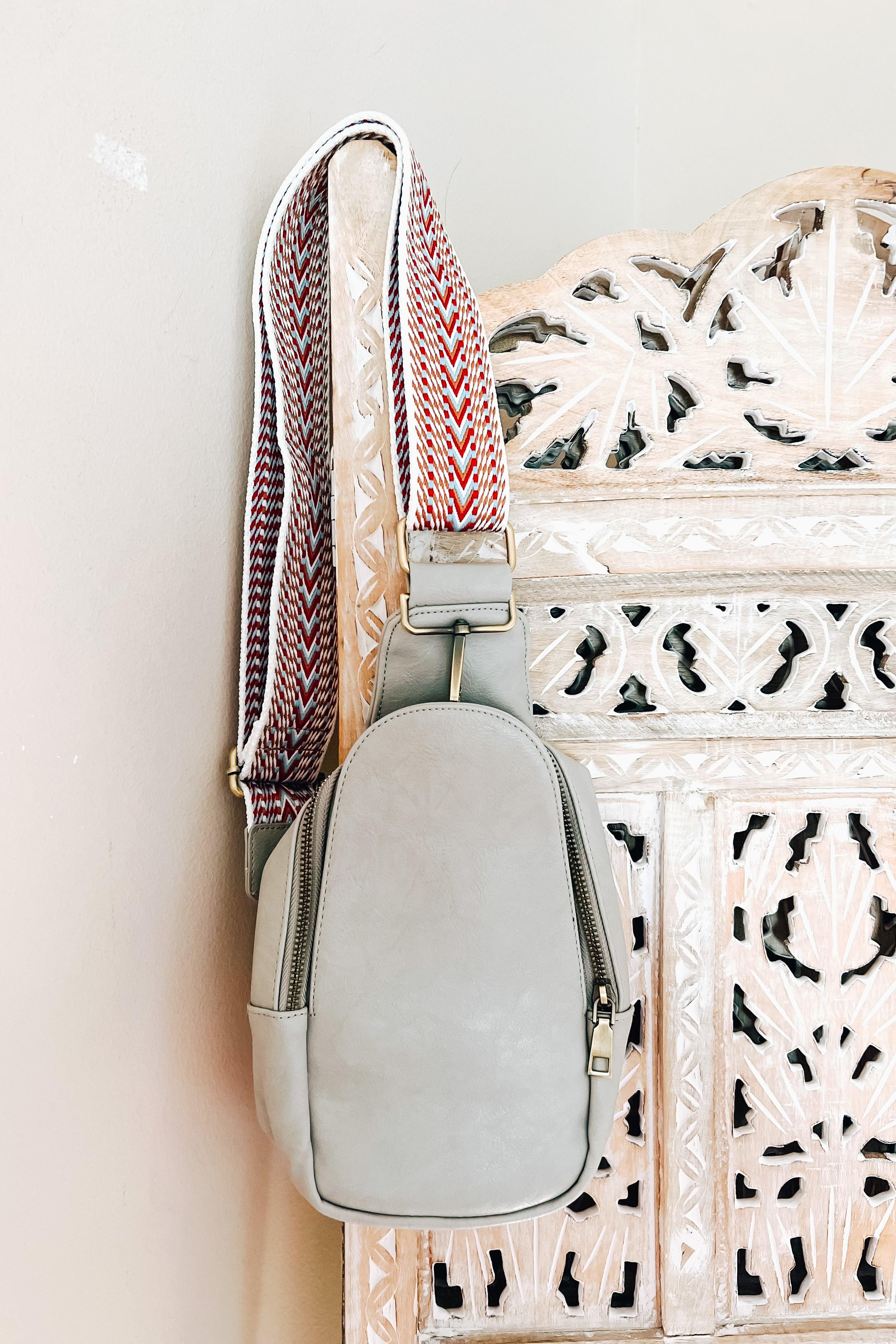 Grey vegan leather sling bag with wide patterned strap hanging on carved wooden panel background