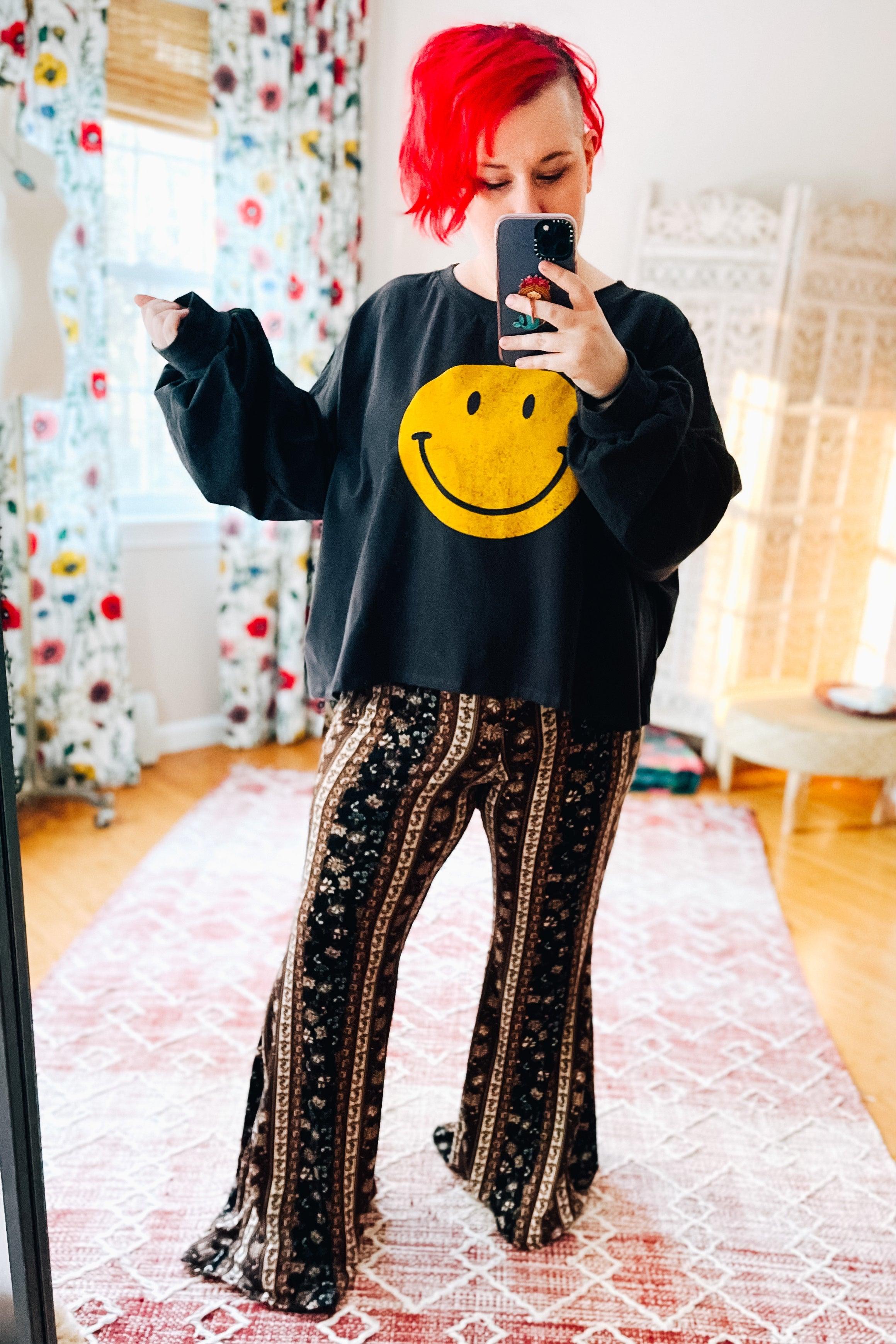 Woman with bright red hair wearing charcoal long sleeve crop top with yellow smiley face and patterned wide-leg pants indoors