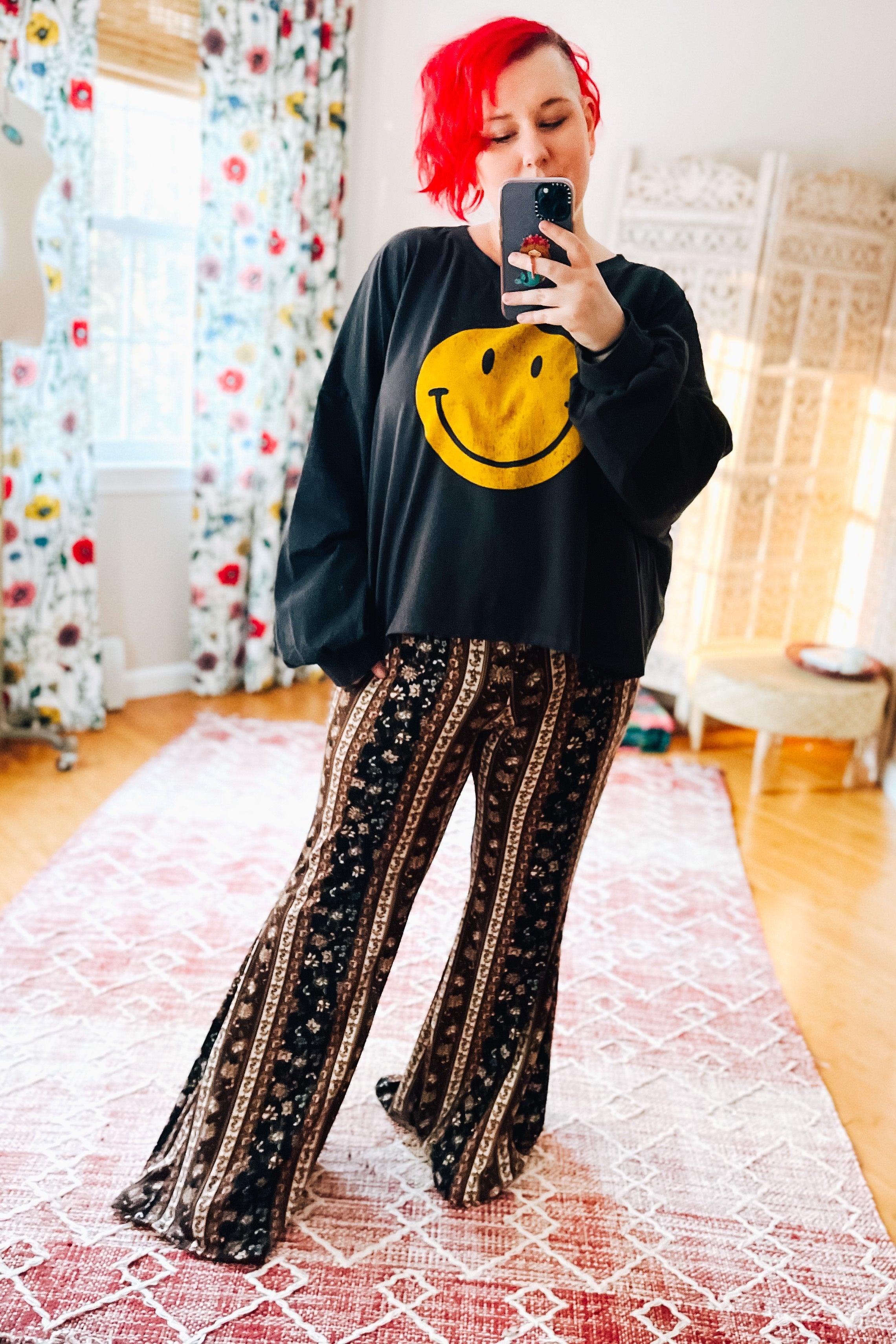 Woman with bright red hair wearing charcoal long sleeve crop top with yellow smiley face and patterned wide-leg pants indoors