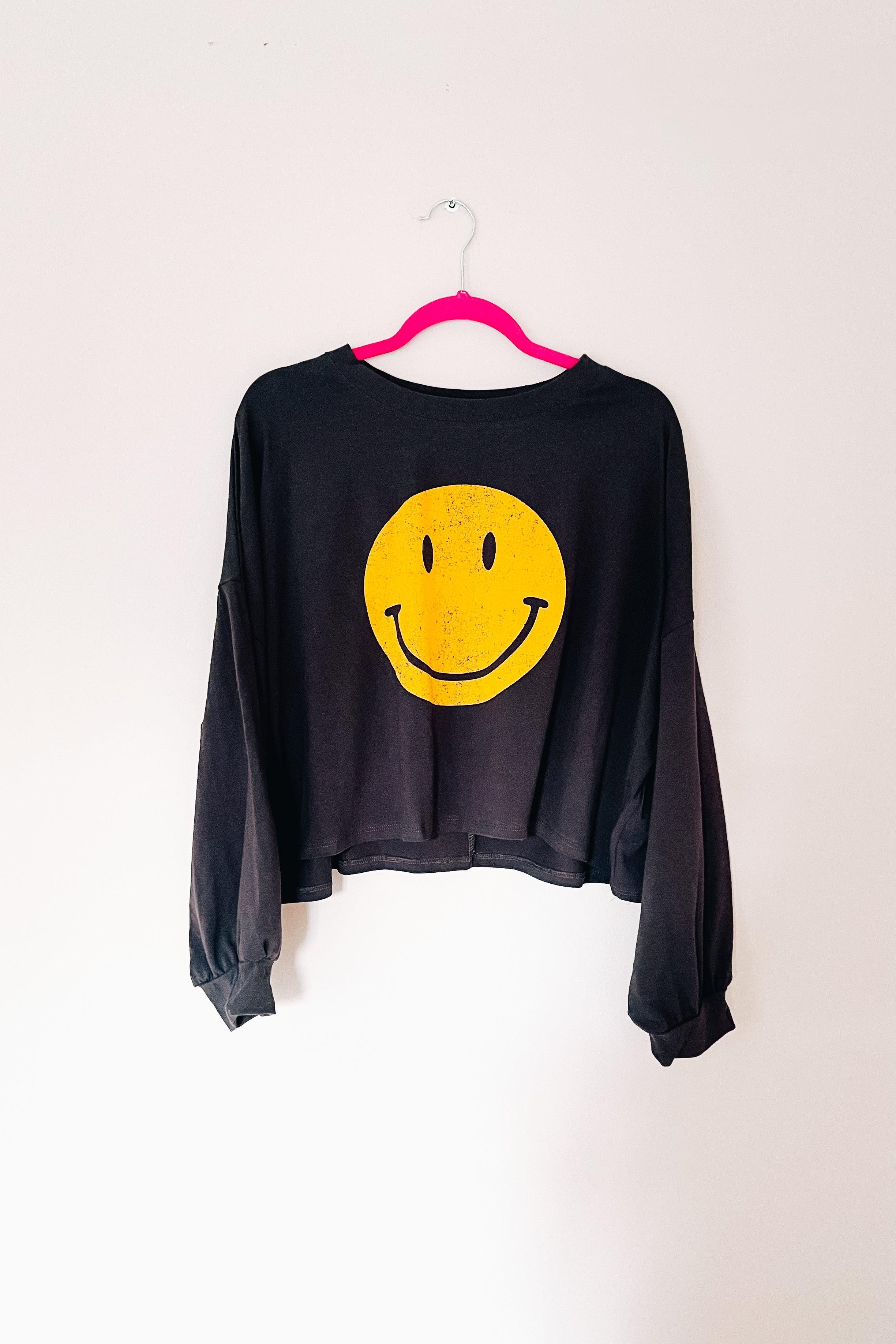 Charcoal long sleeve cropped top with oversized smiley face graphic hanging on bright pink hanger against white wall