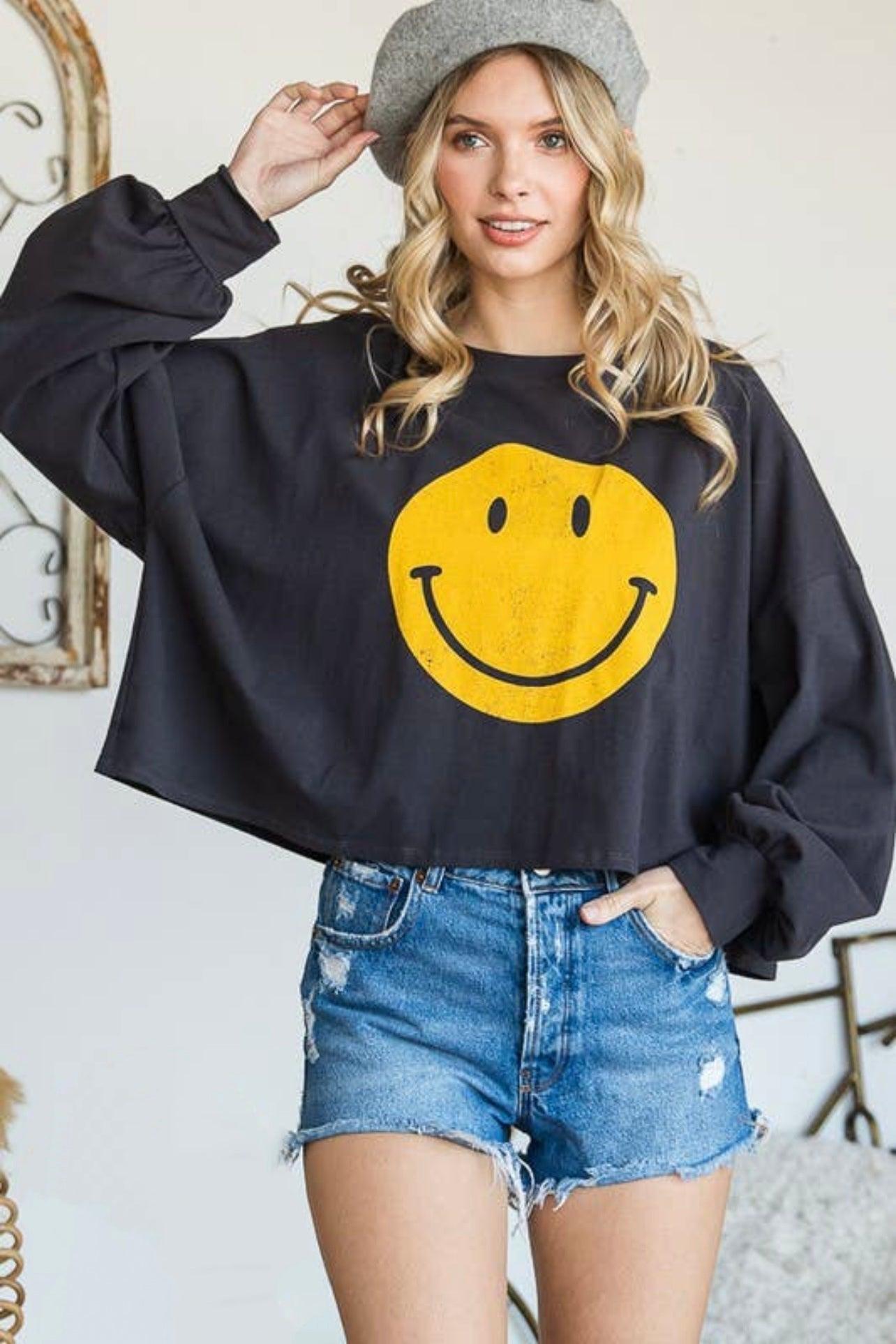 Woman modeling charcoal long sleeve crop top with large yellow smiley face paired with distressed denim shorts and grey beret